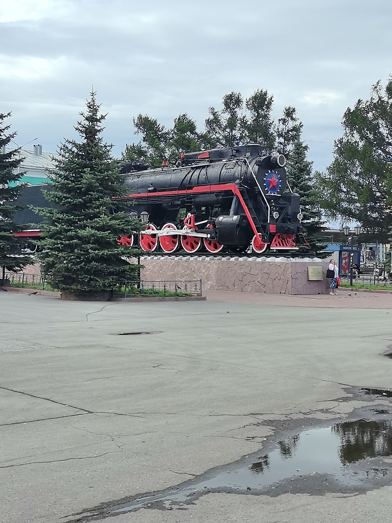 An outdoor, eye-level, medium shot captures a black and red steam locomotive displayed on a stone pedestal. The locomotive is positioned in the center of the frame, with lush green pine trees framing it on either side and a cloudy, overcast sky above. In the foreground, a cracked asphalt ground features several puddles reflecting the surroundings. A few people are visible in the background, near the locomotive, with one woman standing and looking towards it, while others are further back and less distinct. The scene appears to be in a public space, possibly a park or square, given the presence of the monument and public walkways.