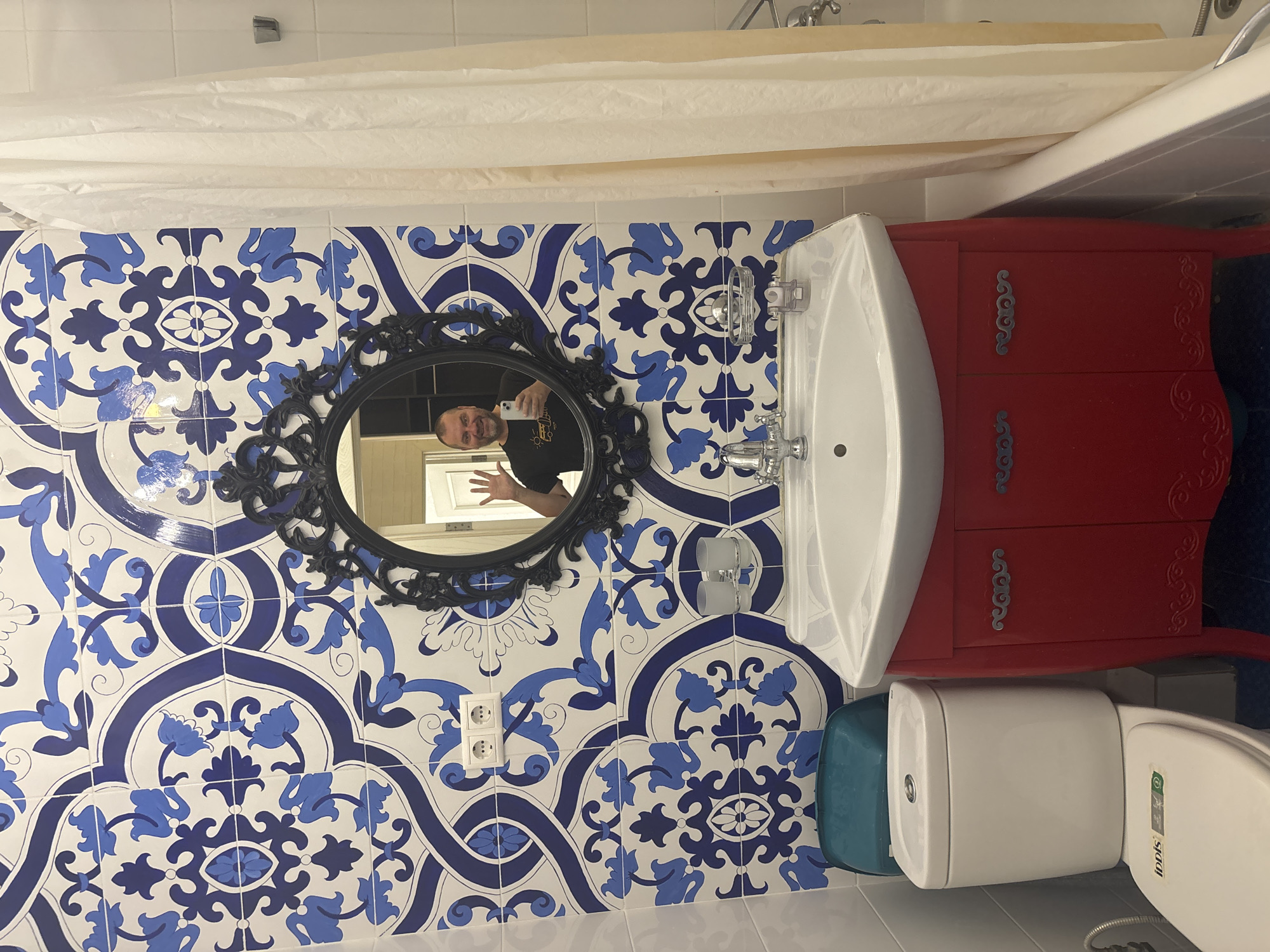 A man is taking a selfie in a bathroom with blue and white tile. He is wearing a black t-shirt and smiling. He is standing next to a toilet and a red vanity with a white sink. A white shower curtain hangs to the right. The bathroom appears to be in a hotel or apartment in Krasnodar, Russia.