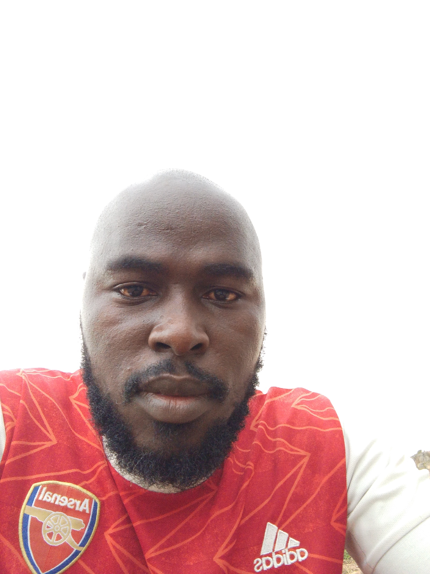 A close-up portrait of a bald, dark-skinned man with a beard, wearing a red Arsenal soccer jersey with white sleeves, taken in Gura Top, Nigeria. The Arsenal emblem is visible on the left side of the jersey and the Adidas logo is at the bottom. The background is washed out.