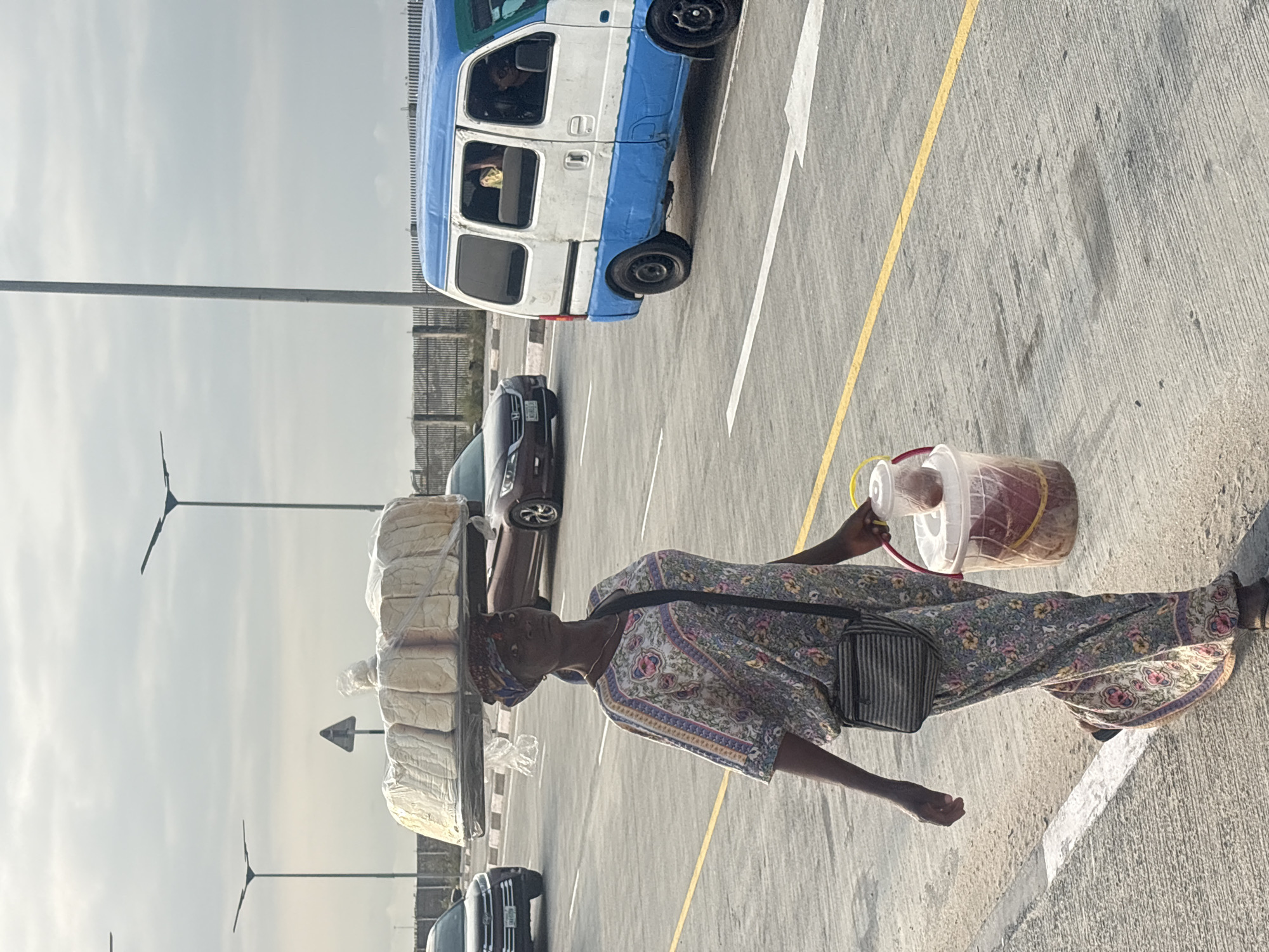 In Itirin, Nigeria, a woman walks along a paved road, balancing a large stack of bread wrapped in plastic on her head. She wears a long, floral-patterned dress and a headscarf. A black crossbody bag with a striped pattern is slung across her body. In her right hand, she carries a bucket with a yellow handle, containing some red liquid, and a smaller plastic container on top. The road has white and yellow lane markings. A blue and white van is visible on the right side of the road, and other vehicles are in the background, including a dark-colored SUV. The sky is overcast, and streetlights are present along the road.