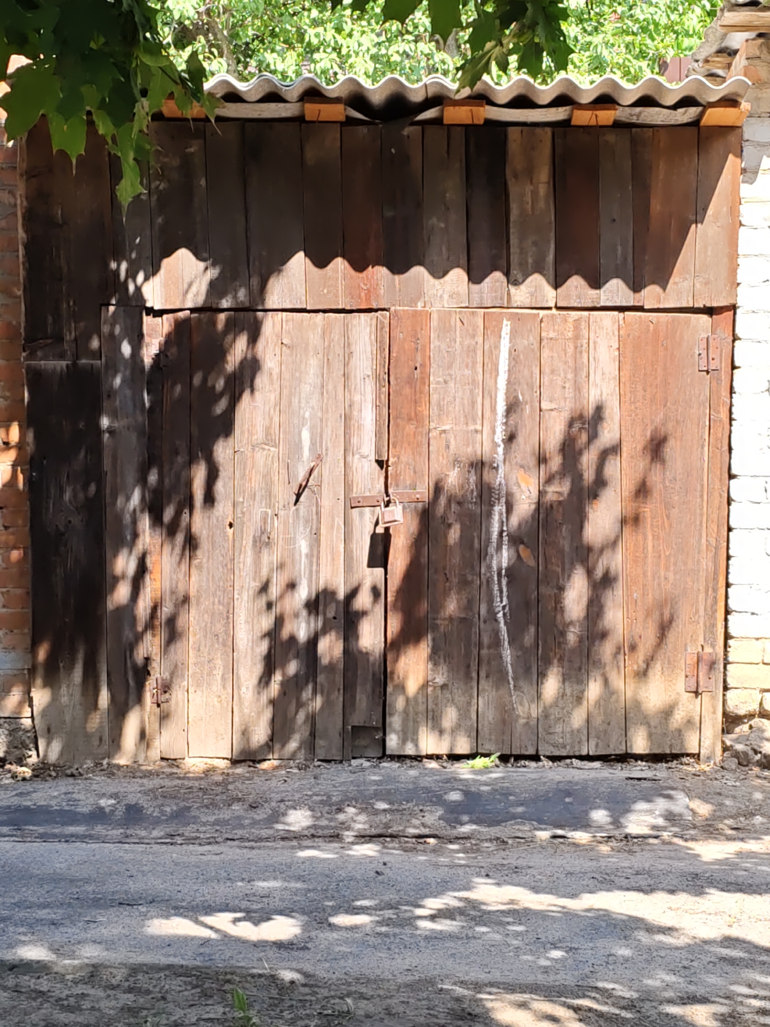An outdoor shot shows a rustic wooden gate that is closed. The gate appears old and weathered, with vertical planks of varying shades of brown. A padlock is attached to the gate in the center. The gate is partially covered by tree branches and leaves, and shadows of the foliage are cast across its surface. The gate is flanked by brick walls, one of which is covered in white brick. Above the gate, a corrugated roof made of what appears to be slate or asbestos material is visible, supported by small wooden beams. The ground in front of the gate is paved with asphalt, and more leaf shadows are visible on it. The scene suggests a rural or older urban setting, possibly in a backyard or alleyway.