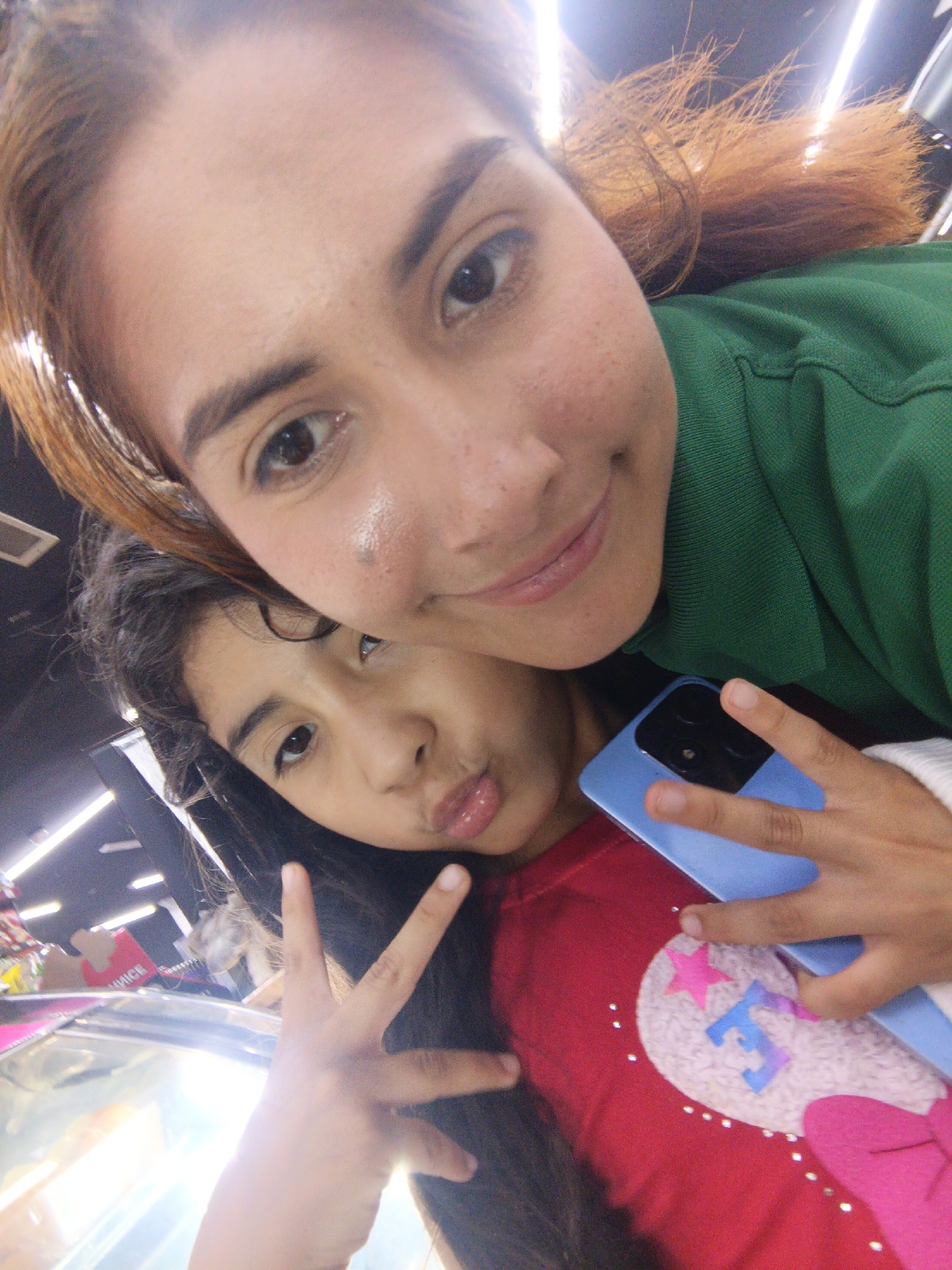 Two females are shown in a close-up, selfie-style photo. The female in the background is an adult with tan skin, brown hair with red highlights, and is wearing a green shirt. The female in the foreground is a child with long dark hair. She is wearing a red shirt with a white heart-shaped design, a pink star, and a blue "E" on the front. She is making a peace sign with her left hand. She is holding a blue cell phone. The background is blurry, but appears to be a commercial location. There are bright lights on the ceiling. The location is presumed to be Altagracia, Venezuela.