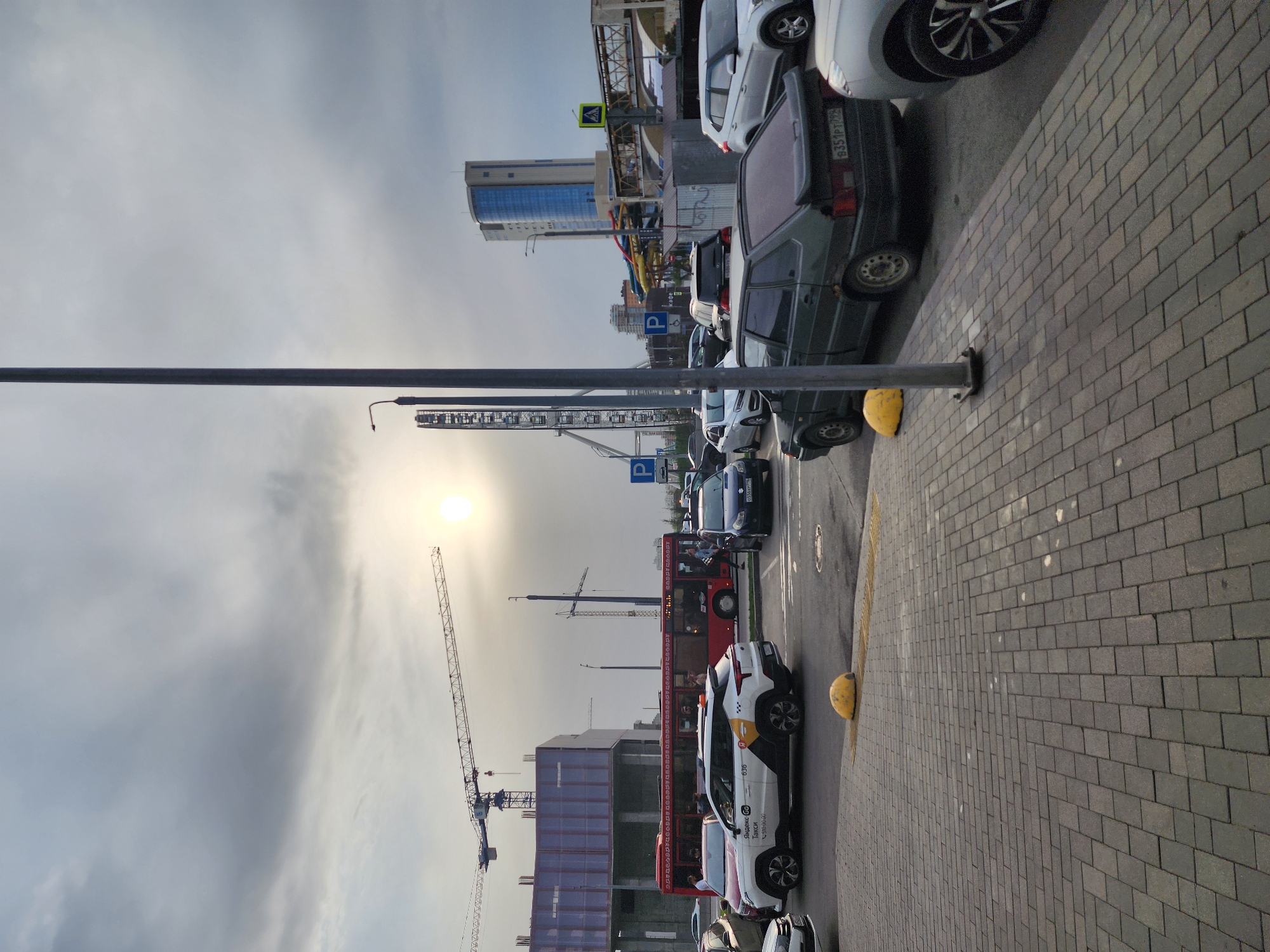 The image depicts a street scene in Kazan, Russia. The foreground features a paved walkway with yellow bollards separating it from a parking area. Cars are parked along the parking area. A red double-decker tourist bus is visible, along with several smaller vehicles. In the background, there are buildings, including a high-rise with blue glass panels, and a construction site with a crane. The sky is overcast with the sun partially visible. A tall pole is visible in the middle of the picture.