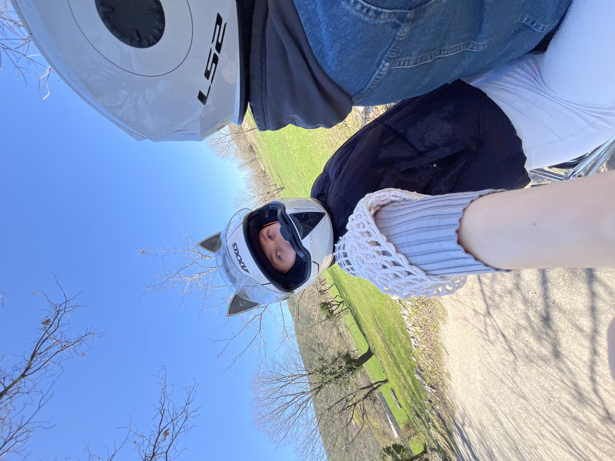 A sideways shot depicts two people wearing helmets riding a motorcycle on a path lined with green grass and trees, likely near Kovači, Montenegro. The first rider wears a white helmet, a grey jacket, and jeans. The second person wears a helmet with cat ears and a light-colored top with a white crocheted shawl. The path appears to be concrete, with shadows cast by the nearby trees. The sky is clear and blue.