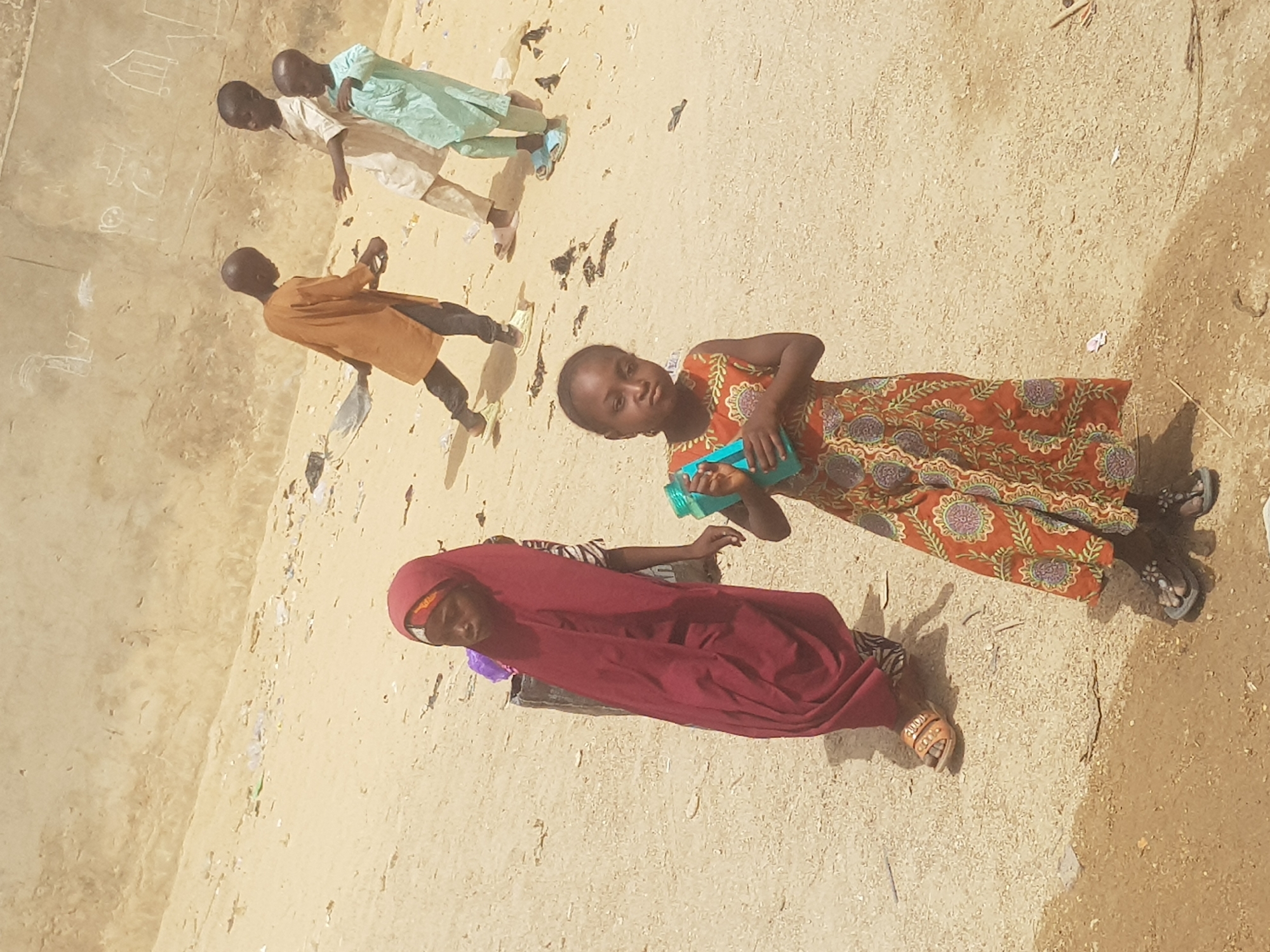 The image depicts a group of children and a woman in an outdoor setting, possibly a schoolyard, in Dutsinma B, Nigeria. The ground appears to be sandy and dusty, with some scattered debris. A wall with chalk drawings is visible in the background.
Four children are seen standing near the wall, dressed in traditional clothing. A young girl in an orange patterned dress is standing in the foreground, holding a blue container. A woman wearing a red headscarf and dress is also present. The people are interacting casually, with the children seemingly engaged in play or conversation.