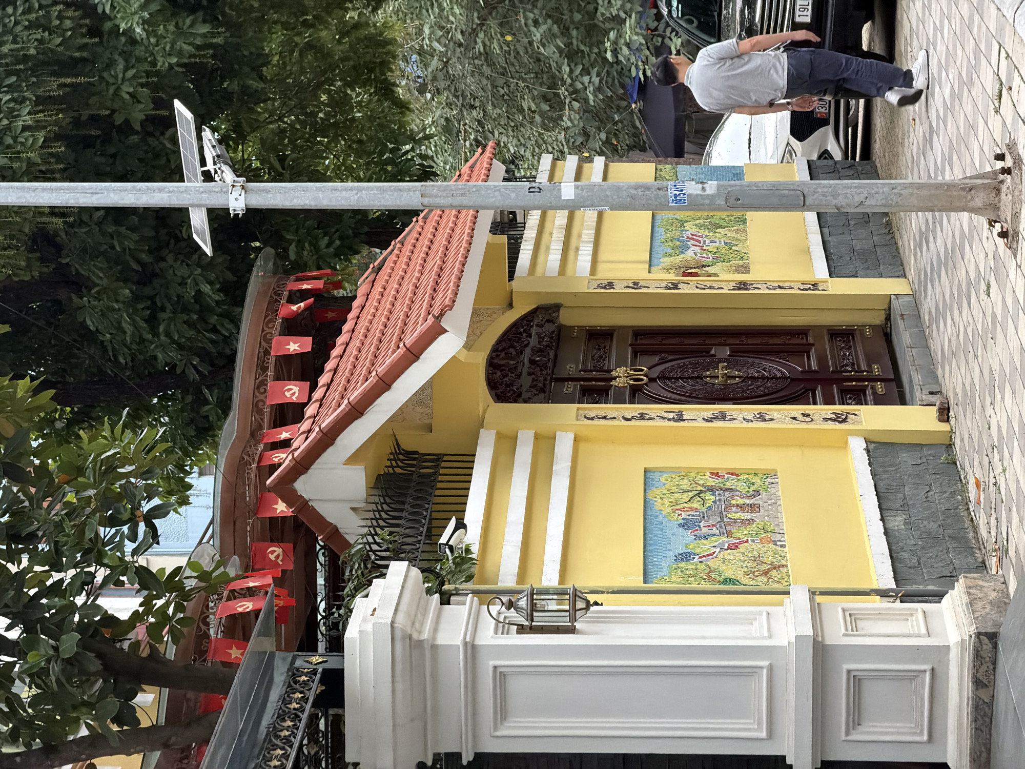 The image depicts a street scene in Hanoi, Vietnam. A man in a gray polo shirt and blue jeans walks away from the camera on a tiled sidewalk. He is in front of a yellow wall with red-tiled roofing and a dark wooden gate. The wall features mosaic art and decorative horizontal white stripes. Red flags with yellow stars and communist symbols hang from an awning above the wall. A street light with a solar panel stands on the sidewalk to the right of the gate. A parked black car is visible to the right of the man. The environment suggests a residential or commercial area within the city.