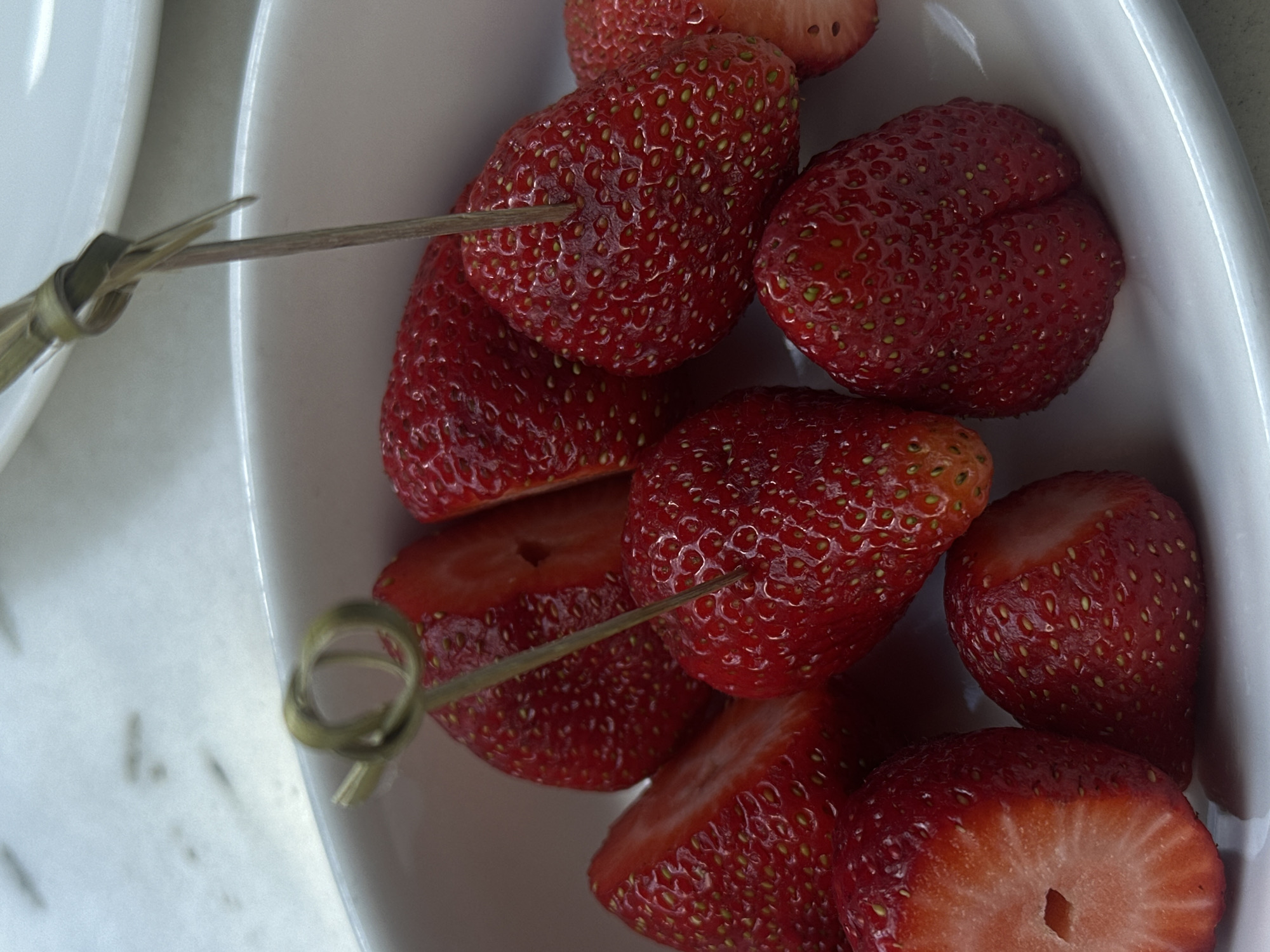 The image shows a close-up of a white bowl filled with several cut and whole strawberries. Two small, decorative wooden skewers are inserted into some of the strawberries. The scene appears to be indoors, possibly on a table. There is no evidence to suggest that this image was taken in Yerevan, Armenia.