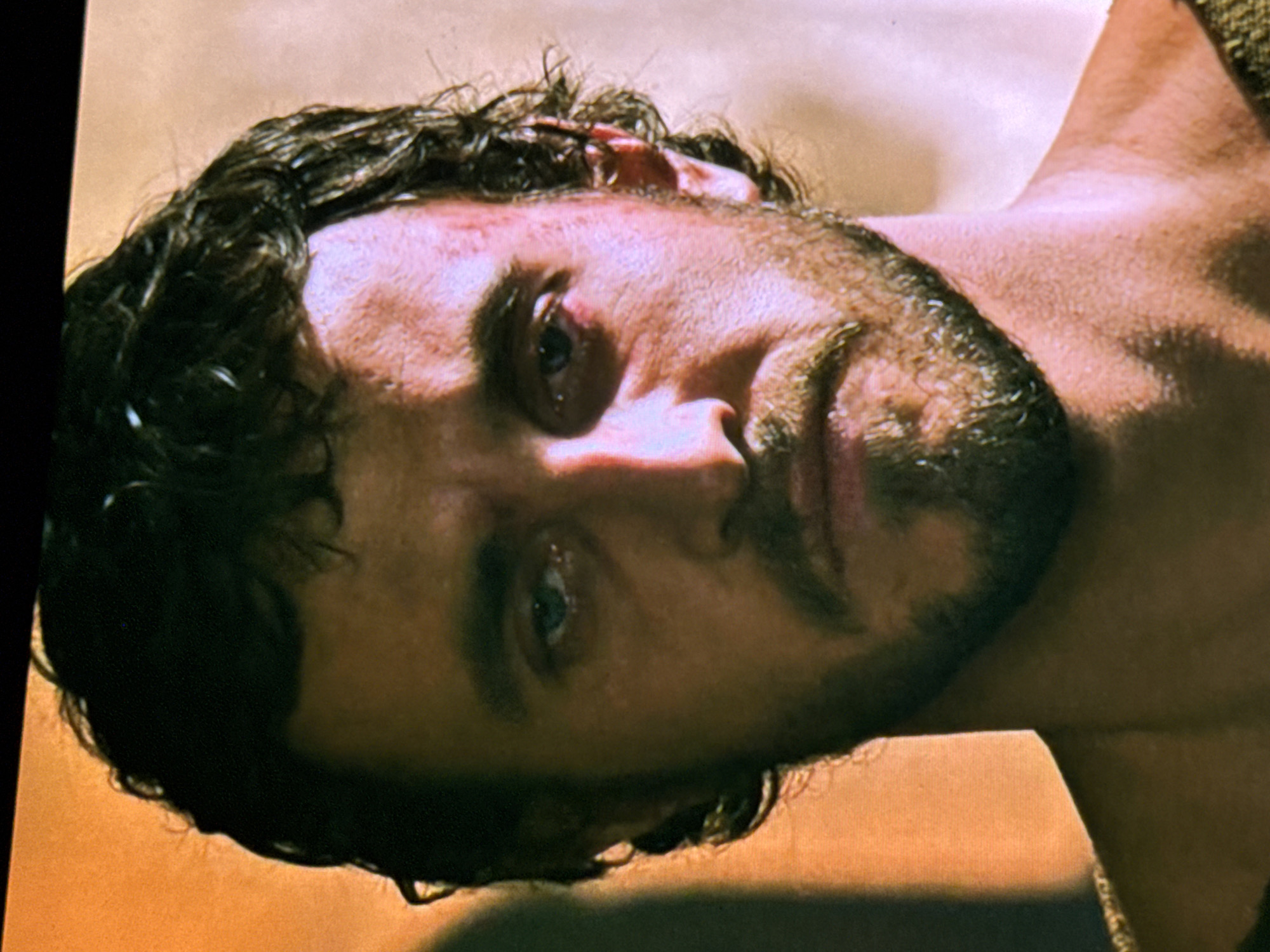 The image displays a close-up portrait of an adult male, from the shoulders up, facing directly towards the viewer. The man has dark, curly, and slightly disheveled hair, appearing damp. He has a short, dark beard and mustache covering his chin, jawline, and upper lip. His eyes are blue-grey and appear to be red-rimmed and moist, suggesting distress or fatigue. The skin on his face is somewhat ruddy, with minor marks or abrasions visible on his forehead and around the eye area. His mouth is closed, with a subtle downturn, contributing to an expression of solemnity or sadness. Visible on his right shoulder is a portion of what appears to be a textured, possibly woven, garment in an olive-green or earthy brown color. The lighting is warm and directional, creating shadows and highlights that emphasize facial contours. The background is out-of-focus and consists of warm, ambiguous tones: a darker orange-brown on the left and a lighter, pale beige or off-white on the right, providing no discernible architectural or environmental details. The image contains no visual information, such as landmarks or identifiers, that would indicate a location in the city of Sofia, Bulgaria, or any other specific geographic place.