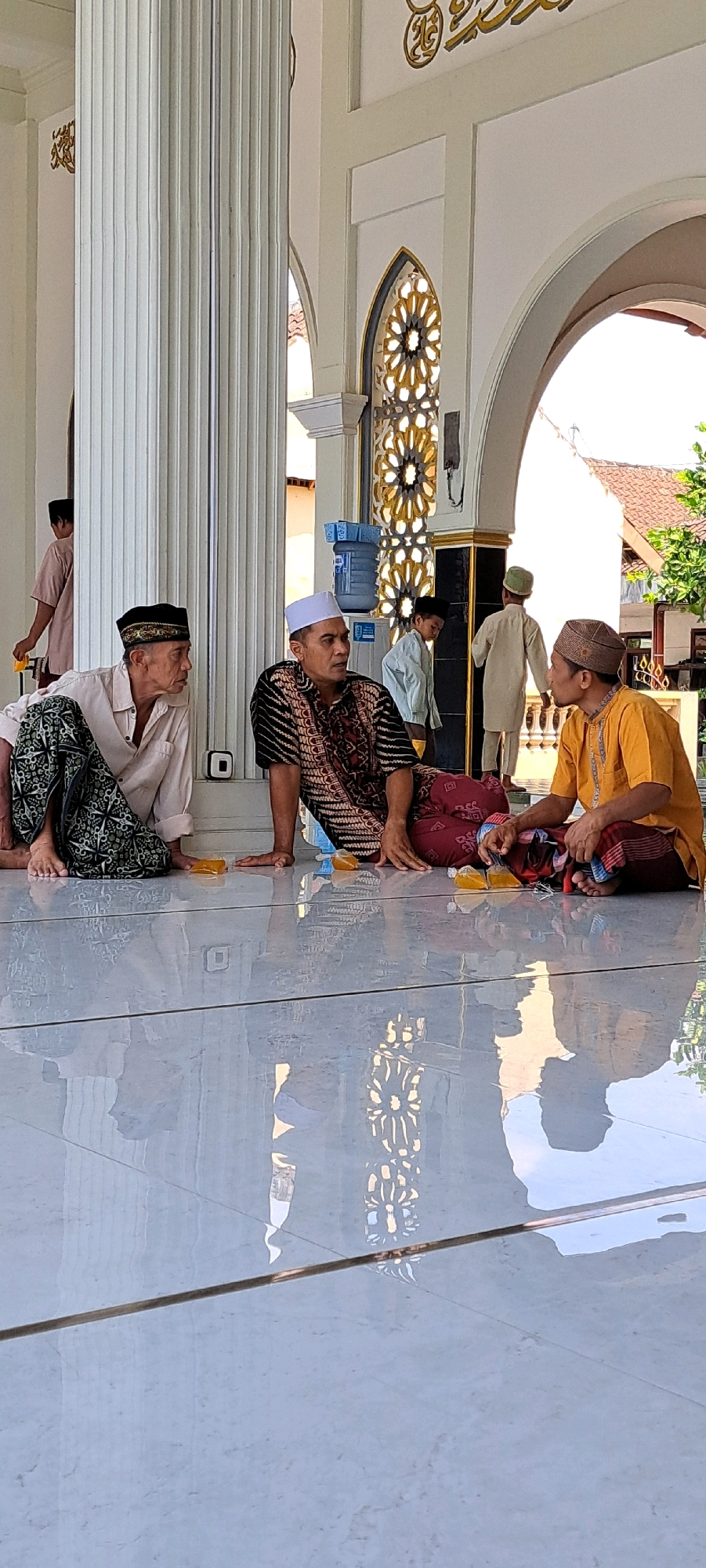 In the city of Sumber Gedang, Indonesia, three adult men are seated on a reflective, tiled floor, appearing to be in conversation. The man on the left is wearing a light-colored shirt, a black cap, and a patterned sarong. He is looking to his right. The man in the middle is wearing a white cap, a patterned shirt with black, red, and gold elements, and a red sarong. He is looking towards his right. The man on the right is wearing a patterned cap and a yellow shirt with a red and blue sarong. He is looking towards his left and gestures with his hands. Yellow packets are visible on the floor in front of the men. Behind them, there are architectural elements including fluted columns, arches, and decorative window grilles. Water coolers are also present. In the background, other individuals, including children, are visible walking or standing. The scene appears to be an outdoor covered area, possibly a mosque or community building, given the attire and setting.