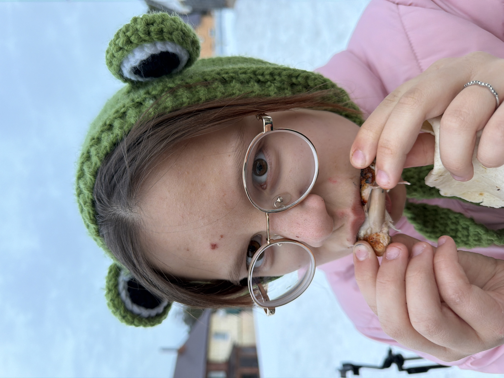 A young person is depicted outdoors in Chaikovsky, Russia, during what appears to be winter, consuming food. The subject wears a distinctive green crocheted hat with frog-like ear protrusions and a pink puffer jacket. Round, gold-rimmed spectacles are worn, and medium-brown hair frames the face, which exhibits a reddish blemish on the forehead. The individual holds a piece of cooked meat on a bone to their mouth with the right hand, while the left hand holds a section of flatbread. A silver-colored ring with a beaded texture is visible on the ring finger of the left hand. The background shows a snow-covered ground and blurry residential buildings, one with a reddish roof, under an overcast sky. A dark, out-of-focus object, possibly a tripod or stand, is partially visible on the far left.