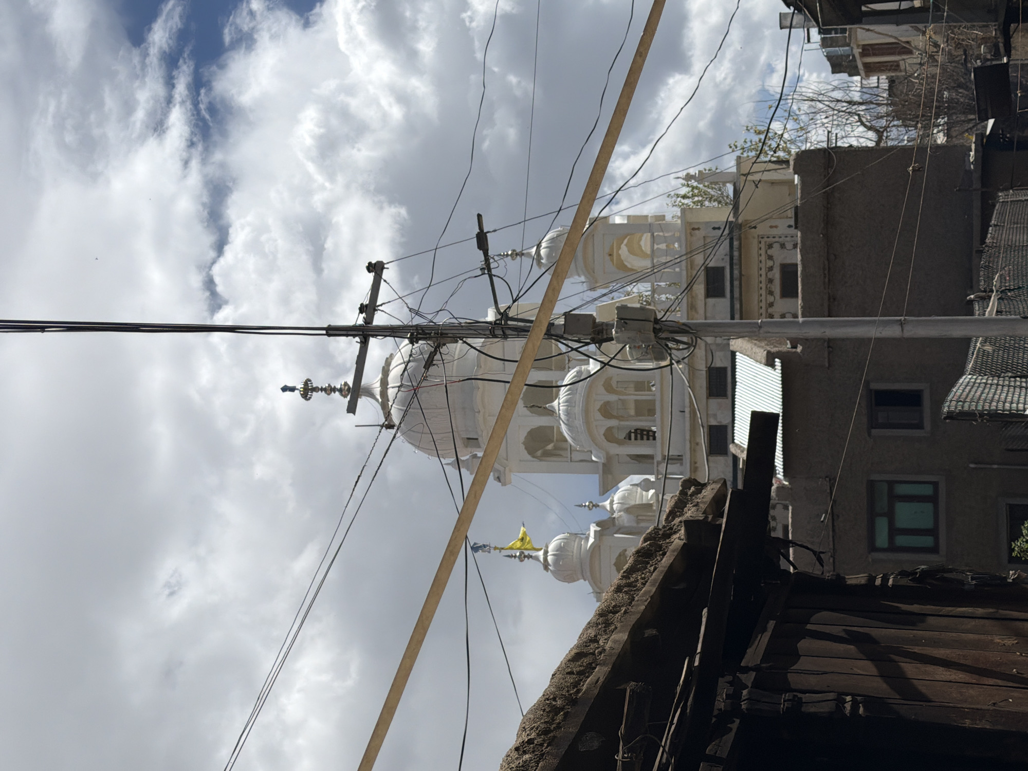 The image shows a white building with domes and a yellow flag, likely a Sikh temple, seen through a tangle of power lines in Leh, India. In the foreground, a dark wooden structure with a rough roof juts into the frame on the left. Behind the wooden structure and the power lines, the multi-domed white building stands against a cloudy sky. A utility pole with numerous wires is prominently positioned in the center, partially obscuring the building. To the right of the pole, more of the white building is visible, along with other structures and bare trees. The overall scene depicts an urban environment with both traditional architecture and modern infrastructure.