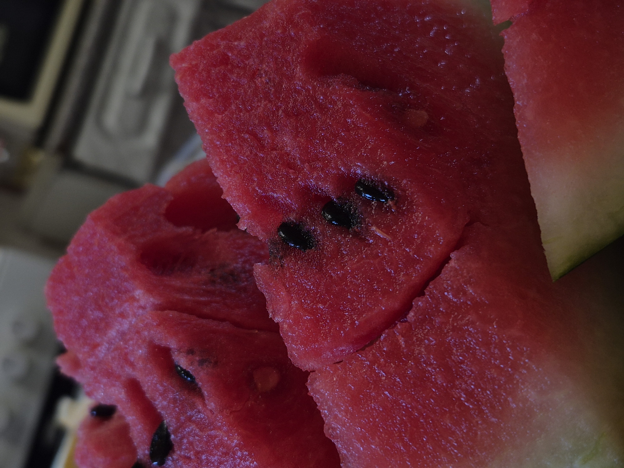 A close-up, low-angle, indoor shot displays several pieces of cut watermelon. The watermelon is a vibrant red with visible black seeds embedded in the flesh. The texture of the watermelon appears slightly granular and moist. In the background, a blurred kitchen environment is suggested by muted tones and indistinct shapes, possibly indicating countertops or appliances. The scene is likely located within a residential kitchen in the city of Дойная, Russia, given the context provided. No people are present in the image, and the focus is entirely on the fruit.