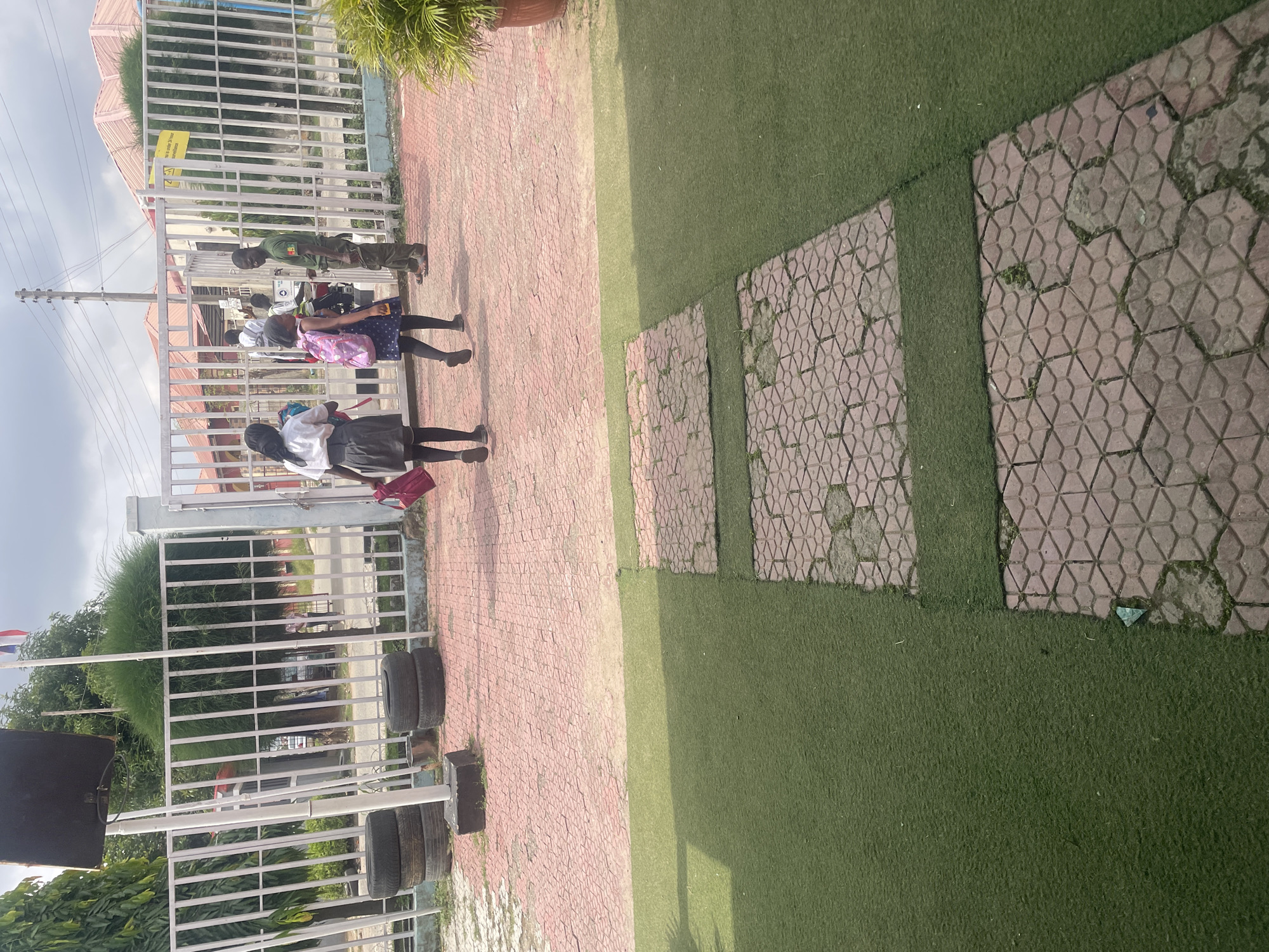 The image shows the entrance to a school in Abuja, Nigeria. Three young girls, likely students, are walking towards a gate. Two of the girls are carrying backpacks, and one is carrying a pink handbag. They are dressed in school uniforms. A male security guard, wearing a green uniform, is standing at the gate, facing the girls and appearing to interact with them. The ground in front of the gate is paved with red interlocking bricks, and there is artificial turf on either side of a stone pathway leading away from the gate. The background shows the school buildings, fences, and trees. The sky is overcast.