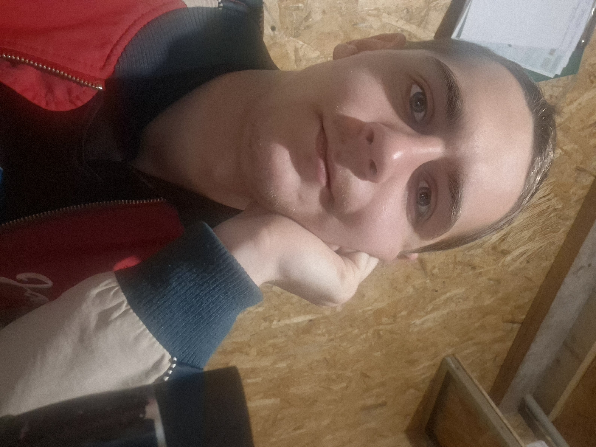 A person is resting their head on their hand while posing for a selfie. The person is wearing a red and black jacket with a blue and white cuff. The background consists of wooden paneling. A clipboard with papers is visible in the upper right corner of the image.