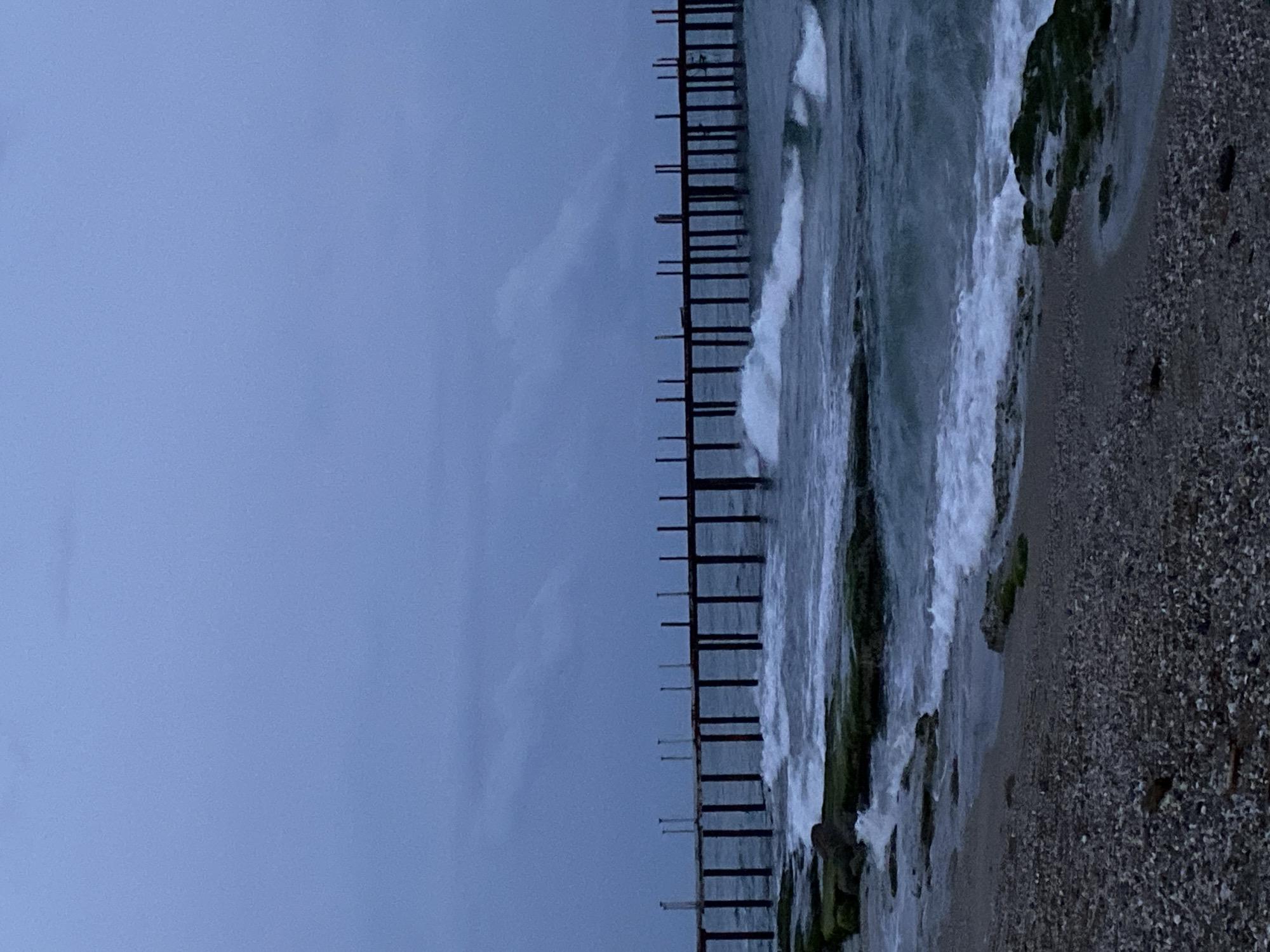 The image depicts a seascape in Alanya, Turkey, under an overcast sky. To the right, a rocky beach transitions into turbulent ocean waves, some breaking against the shoreline. A dilapidated wooden pier stretches into the water, its supports exposed. The beach is composed of pebbles and some algae-covered rocks. The sky is a uniform gray, blending with the horizon. There are no discernible people or other objects within the frame.