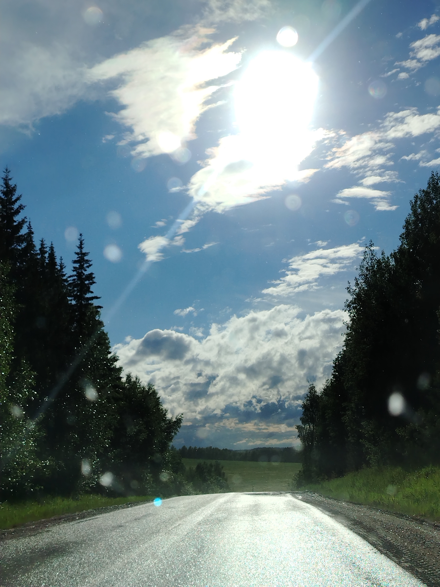 The image shows a road stretching into the distance, flanked by trees on both sides. The sky is visible above, with clouds and a bright sun. The road surface is wet and reflects the sunlight. The location cannot be determined from the image.