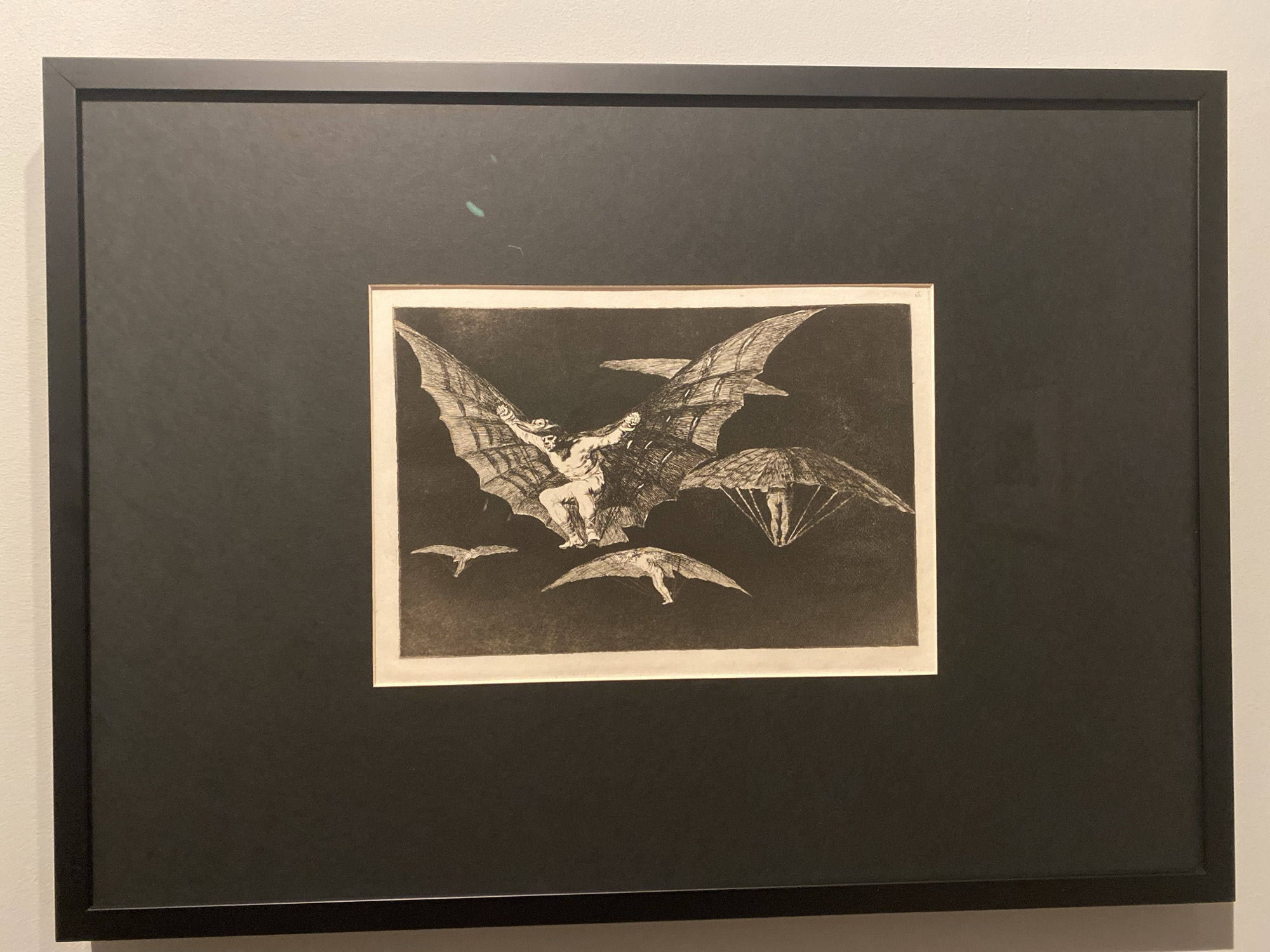 The image displays a framed artwork, an etching titled "The Sleep of Reason Produces Monsters" by Francisco Goya. The artwork depicts a scene with a large winged creature, possibly a bat or a winged humanoid, carrying a man. Below the main creature, there are other figures in flight, one appearing to use a parasol-like device and another with outstretched wings. The overall impression is one of a surreal and nightmarish landscape. The artwork is matted in black and housed in a simple black frame. The background of the image is a plain white wall. There are two faint teal smudges on the matting above the artwork. Based on the context of the artwork, it is a depiction of a print rather than a photograph of a scene in Sofia, Bulgaria.