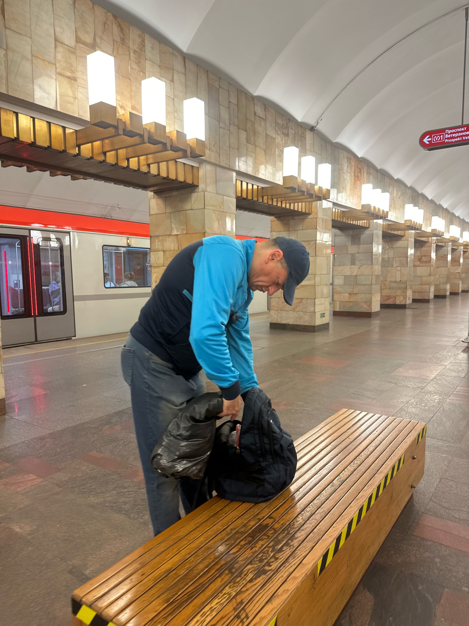 A man wearing a blue jacket and jeans is seen on a subway platform in Murino, Russia. He is bent over a wooden bench, rummaging through a black backpack. The subway train is visible on the left side of the image, with its doors open. The station features stone walls and a white arched ceiling with overhead lighting. A red directional sign with white text indicating "Prospekt Veteranov" (Veteranov Avenue) is also visible. The platform floor is made of stone tiles.