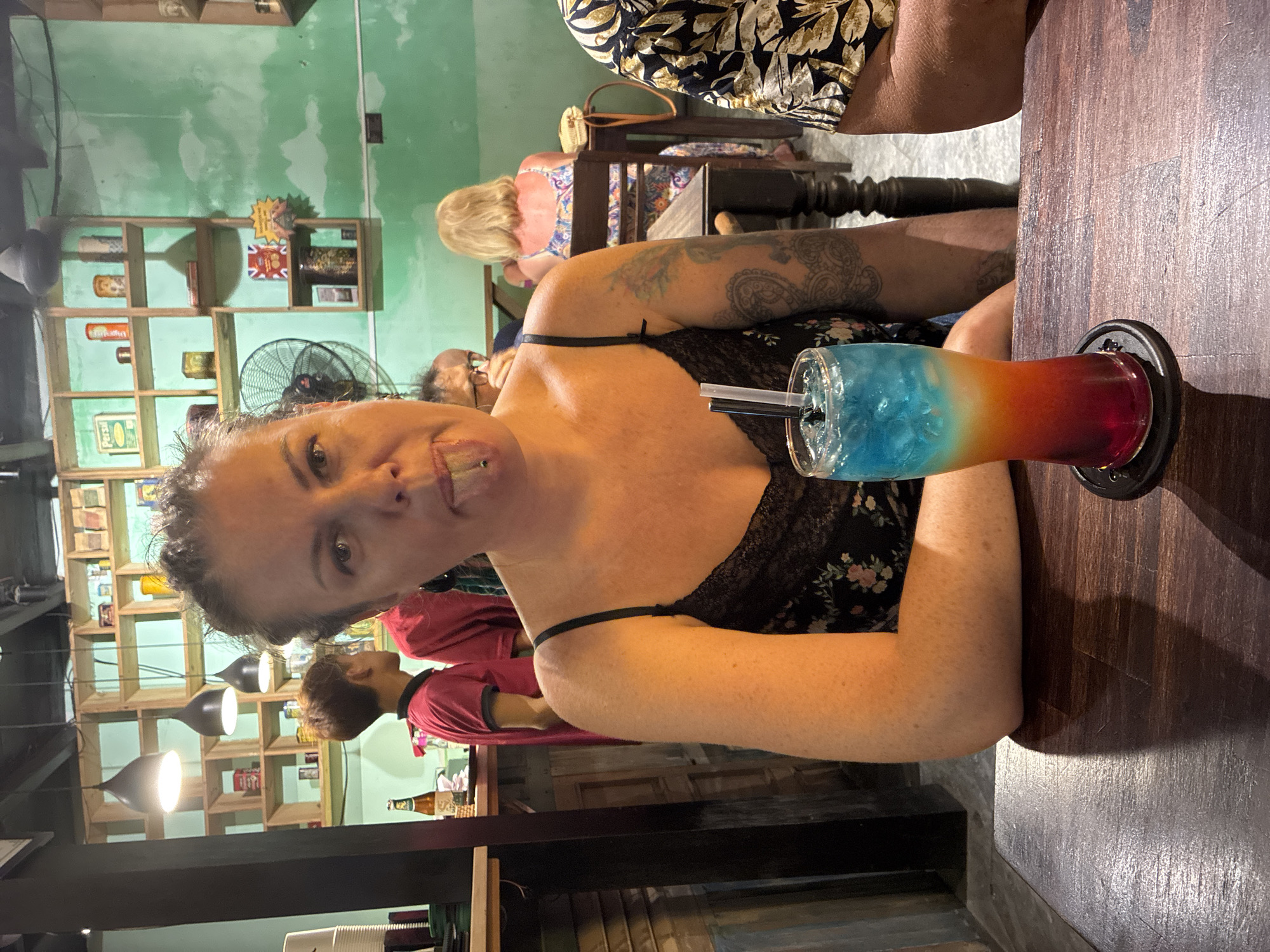 The photograph, taken at an unidentified location within Hội An Ward, Vietnam, shows a woman seated at a dark wooden table, with a layered cocktail of blue, orange, and red hues placed to her right. She is wearing a floral patterned top with lace detail and black straps, and has a visible tattoo on her right arm. Other individuals are present in the background, including a fair-haired woman in a patterned top and a person in a red shirt. The environment includes shelving units stocked with various items and several hanging light fixtures. The walls are painted a light green color. A black fan is positioned above the shelves.
