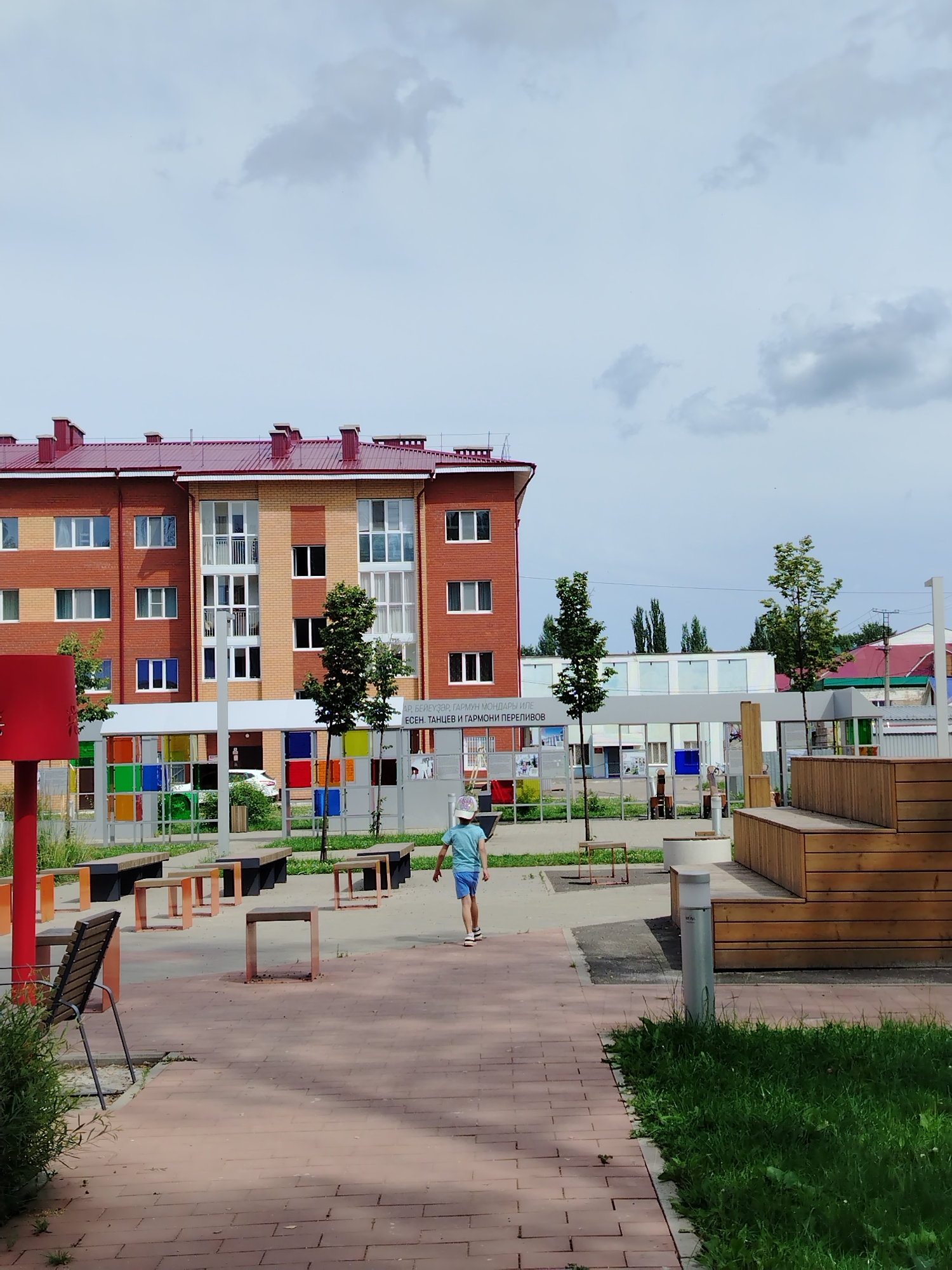 The image depicts an outdoor public space in Yanaul, Russia, featuring a modern residential building with a red roof and brick facade. In the foreground, a child wearing a light-colored hat and blue t-shirt and shorts walks away from the camera on a brick pathway. The area includes several outdoor seating arrangements with benches and tables, some with orange metal legs and others with wooden tops. Adjacent to the building is a fence with colorful stained-glass panels. Beyond this, a wooden tiered seating structure is visible on the right side of the frame. The environment is overcast with a cloudy sky.