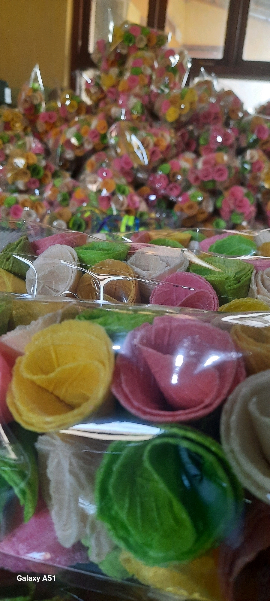 In a close-up, eye-level shot, numerous brightly colored, rose-shaped snacks are neatly arranged and packaged in clear plastic. The snacks, in shades of pink, yellow, green, and white, are layered within transparent containers, creating a visually appealing display. In the background, more of these snacks are visible, also in clear packaging, suggesting a large quantity. The environment appears to be indoors, with soft lighting. The bottom left corner of the image shows the text "Galaxy A51," indicating the device used for capture. The scene is likely a market stall or shop in Jatitengah, Indonesia, where these snacks are being sold.