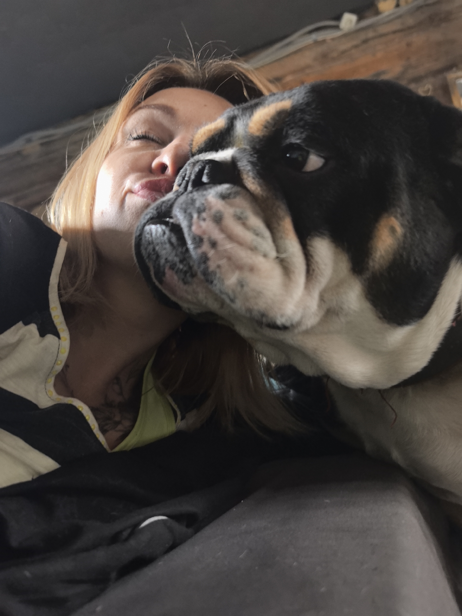 This close-up, angled shot captures a tender moment between a person and a Bulldog indoors. The person, appearing to be a woman with light brown hair, is seen with her eyes closed and lips slightly pursed in an expression of affection, her face resting closely against the dog's head. A Bulldog, with distinctive black, white, and tan markings, is positioned prominently in the foreground, looking slightly to the right with one eye visible. Its characteristic jowls and wrinkles are clearly seen.
The scene conveys a strong sense of warmth and intimacy between the pet owner and their companion. The setting appears to be a cozy indoor environment, likely a home in Noviy, Russia, with soft, diffused lighting suggesting it could be daytime. In the upper background, a wooden beam or structure is partially visible, hinting at the room's interior design. The person is wearing a black top with white and yellow detailing near the collar and a neon green strap of another garment is visible below. A tattoo is faintly visible on the person's neck/collarbone area. Both subjects appear to be resting on or alongside a dark fabric surface, possibly a bed or sofa. No text is visible in the image. The overall mood is one of peace and affection, capturing a loving bond.
