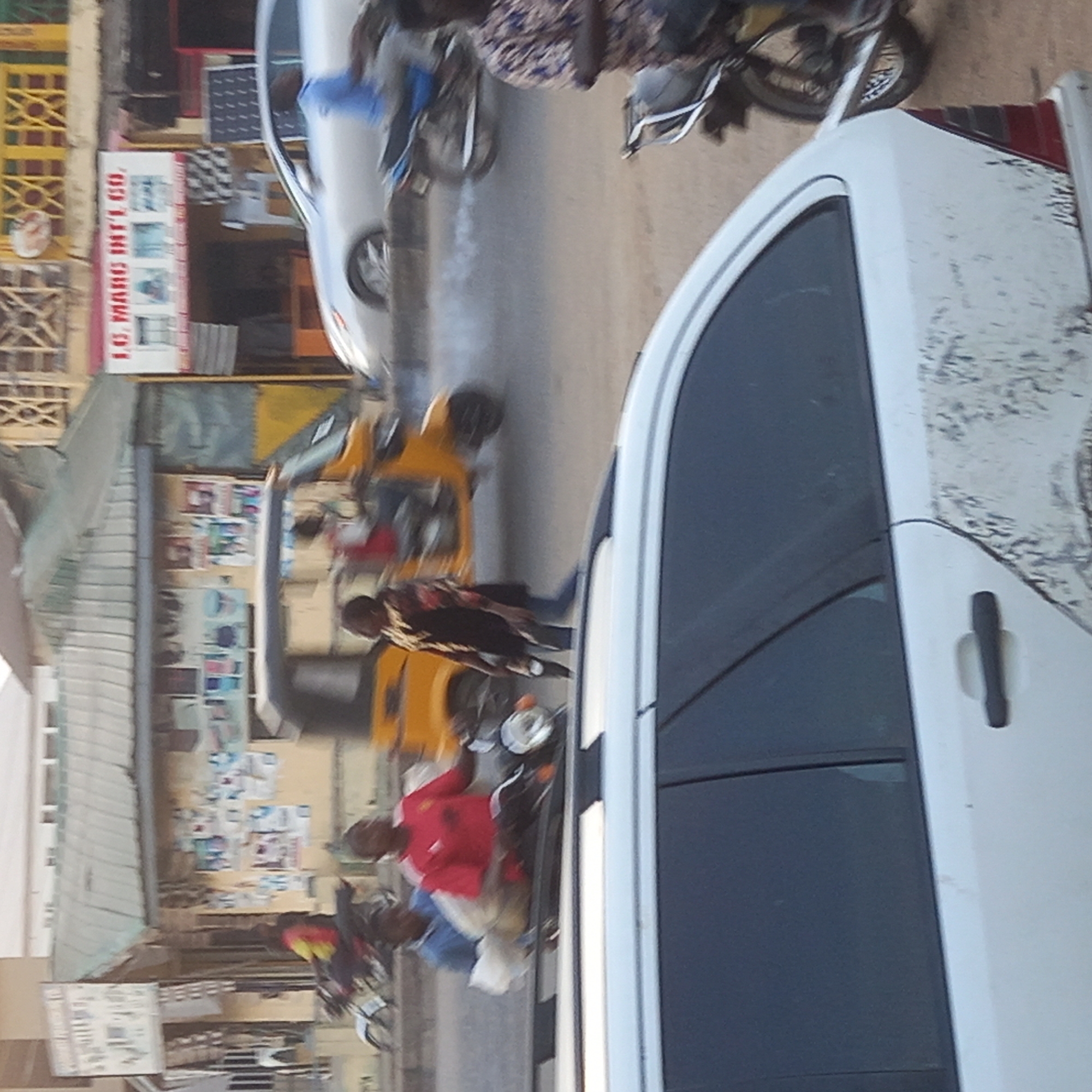 The image depicts a street scene in Sardauna, Nigeria. A white van is prominently featured in the foreground. Behind the van, the street is filled with various vehicles, including a car, a motorcycle, and at least two yellow "keke napep" three-wheeled taxis. People are standing and walking around the vehicles. Buildings line the street, one with a sign that reads "I.O. MAAS INT'L CO.". The buildings are covered in posters and signs.
