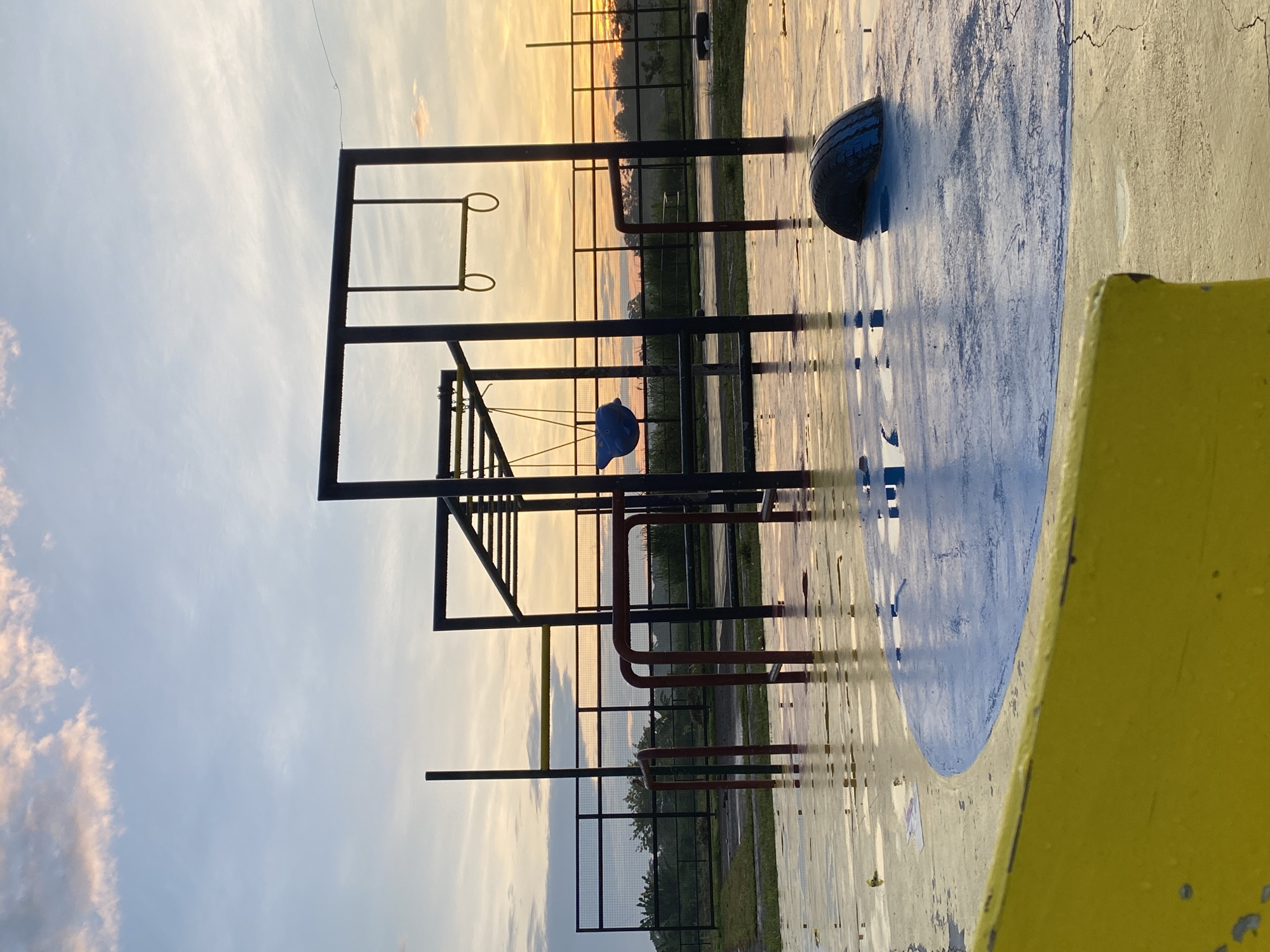 The image depicts an outdoor playground or recreational area at either sunrise or sunset, characterized by warm, golden light across the horizon. The sky, seen at the top, transitions from a soft blue with wispy clouds to vibrant orange and yellow hues near the horizon line.
In the midground, a dark-silhouetted metal playground structure dominates the scene. It features monkey bars, horizontal and vertical supports, and two hanging rings. A distinctive blue, blob-like object, possibly a swing seat or climbing feature, hangs from the structure. Behind this equipment, a chain-link fence runs horizontally, separating the play area from distant greenery.
The ground in the foreground and midground appears to be a paved or concrete surface, noticeably wet and reflective, mirroring the warm colors of the sky. A large, bright blue painted circle or oval is visible on the ground. Towards the bottom right corner, a black tire is partially embedded in the ground, and a bright yellow structure, possibly a curb, bench, or part of another play element, frames the extreme right side of the image.
No people or discernible activities are present, giving the scene a serene and tranquil atmosphere. The reflections on the wet surface suggest recent rain or dew. The golden hour lighting enhances the silhouettes of the structures and creates long reflections. The location context indicates this scene is in Davao City, Philippines. There is no visible text in the image.
