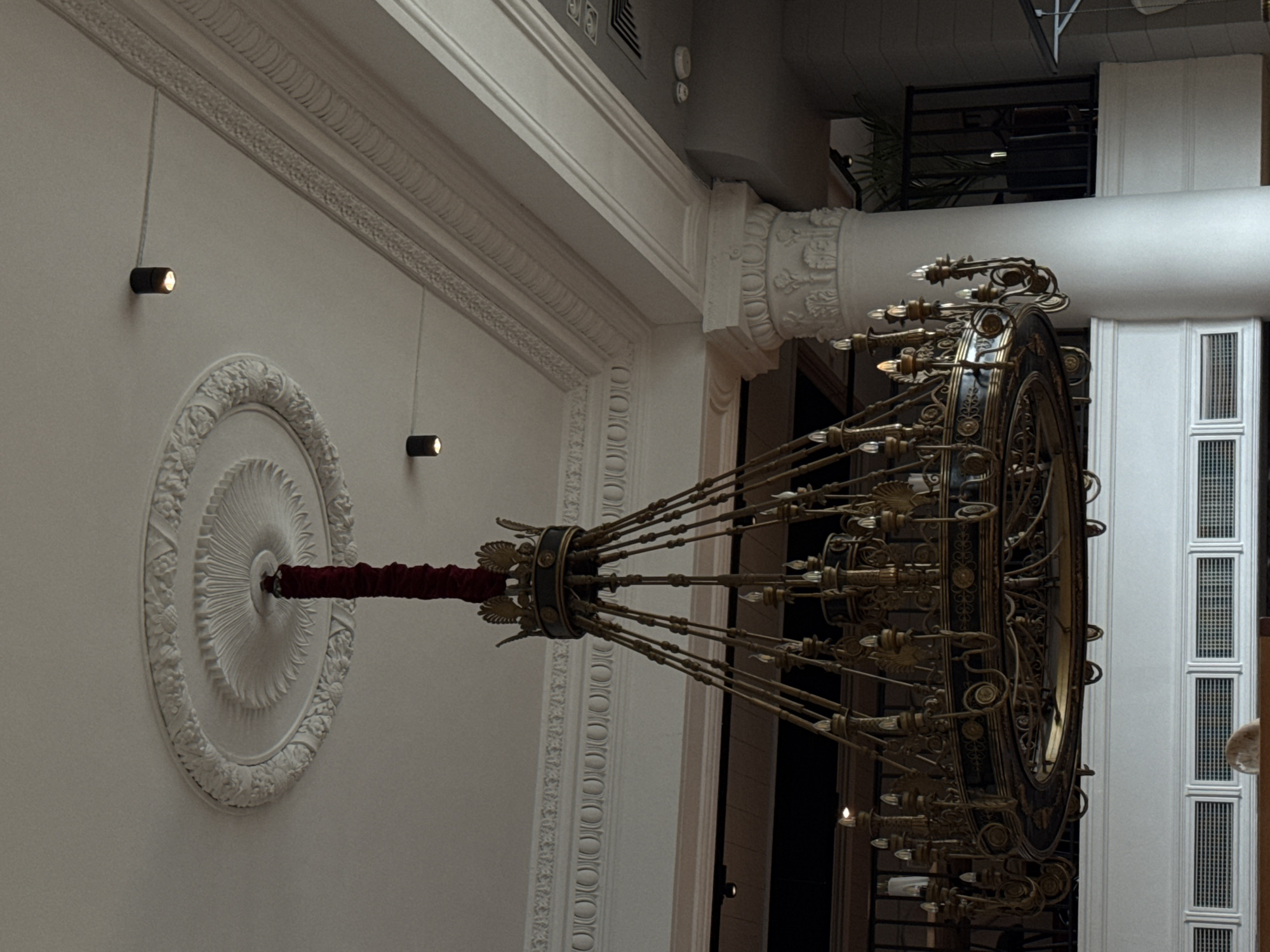 This media file captures a grand interior space, likely a hall or lobby, characterized by elaborate classical architecture. The primary subject is a large, ornate multi-tiered chandelier, suspended vertically from a high ceiling. The chandelier, appearing to be made of dark bronze or brass, features intricate scrollwork, numerous arms, and many small light bulbs, several of which are illuminated. Its central suspension rod is distinctively wrapped in red fabric.
The ceiling, painted a light color, showcases exquisite decorative elements, including a large, circular plaster medallion with a sunburst or petal-like pattern directly around the chandelier's point of attachment. Ornate friezes and moldings run along the ceiling edges and possibly upper wall sections, adding to the classical aesthetic.
In addition to the main chandelier, smaller, modern, black cylindrical spotlights are mounted on the walls, providing focused illumination. A prominent fluted column with an elaborately carved Corinthian-style capital is visible, indicating substantial architectural detailing. Further in the background, a large white structure with vertical panels and grates or vents can be seen, possibly part of a wall, window frame, or ventilation system. A hint of green foliage from a plant is also discernible in the upper background.
The scene appears static, with no activity or people visible. The illumination from the chandelier and spotlights suggests an indoor setting, possibly during the evening or under artificial light conditions, given that some bulbs are glowing. The grandeur and style of the architecture are consistent with significant public or historical buildings found in Moscow, Russia. No visible text is present in any of the images.