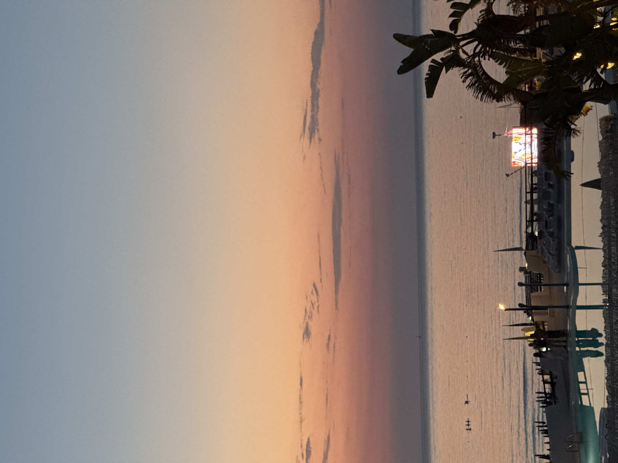 The image depicts a coastal scene at dusk in Isola delle Femmine, Italy. The sky transitions from a pale blue at the top to a soft orange and pink gradient towards the horizon. Several thin, wispy clouds are scattered across the sky. The calm sea reflects the colors of the sunset.
In the foreground, the edge of a swimming pool is visible, with its surface reflecting the surrounding structures and sky. To the right, a large, leafy plant, likely a banana tree or similar tropical foliage, is silhouetted against the sky. Further back, along the shoreline, is a beach setup with lounge chairs, umbrellas, and some structures. A prominent illuminated screen or advertisement is visible amongst the beach furniture, displaying a brightly colored graphic. Several people are standing on the beach, near the pool area, appearing to be in conversation or observing the surroundings. In the distance on the water, a few small boats or buoys are visible. The overall atmosphere is serene, characteristic of a seaside resort at the end of the day.