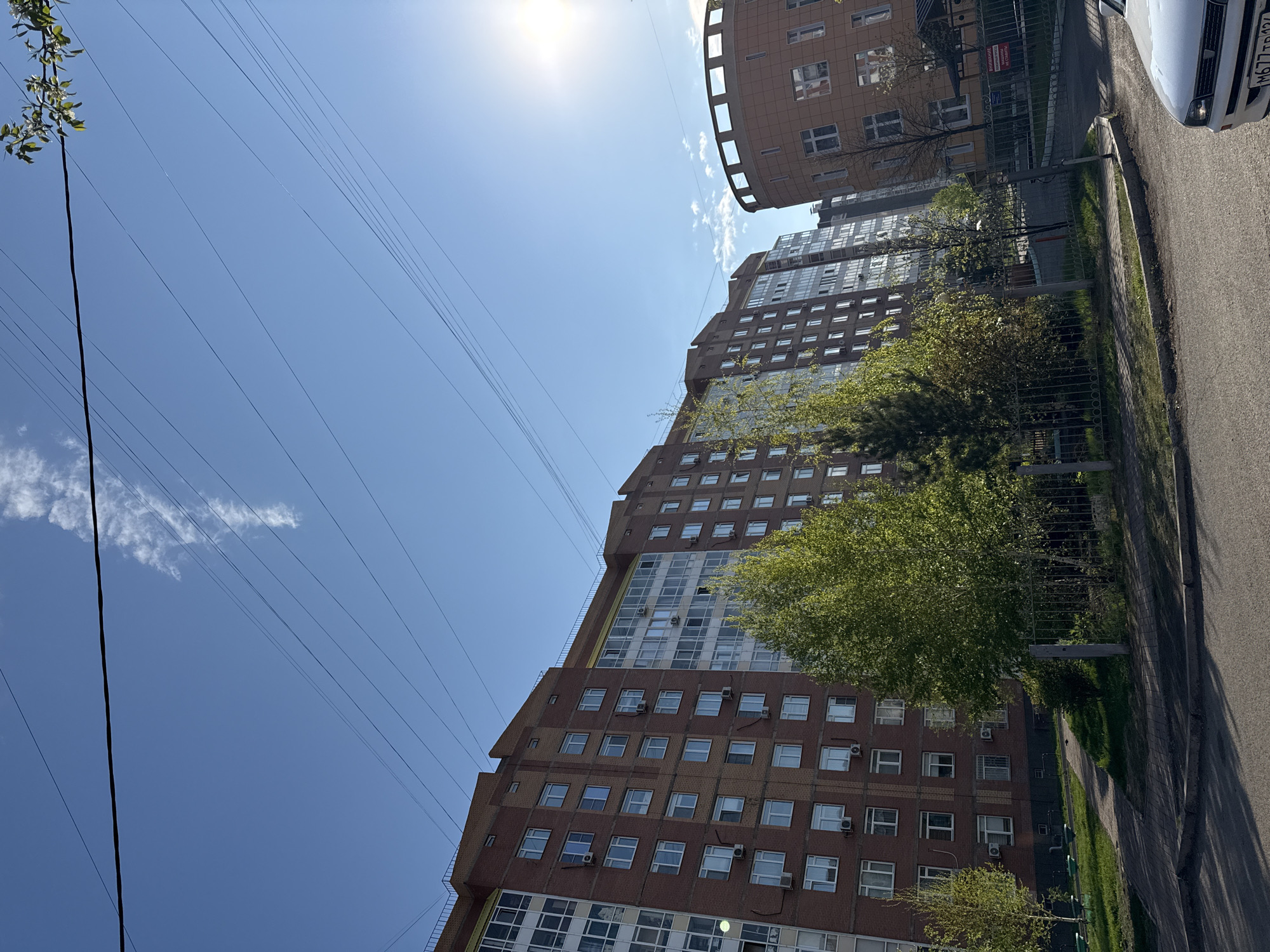 The image shows a tall, multi-story apartment building in Krasnoyarsk, Russia. The building is constructed of reddish-brown brick and features numerous windows and air conditioning units. A rounded structure is visible atop or adjacent to the building. Power lines stretch across the blue sky in the background, partially obscuring the sun. Trees with green foliage are planted along the sidewalk, separating the building from the street. A portion of a white vehicle is visible in the upper right corner of the image. The sidewalk and street are paved with asphalt.