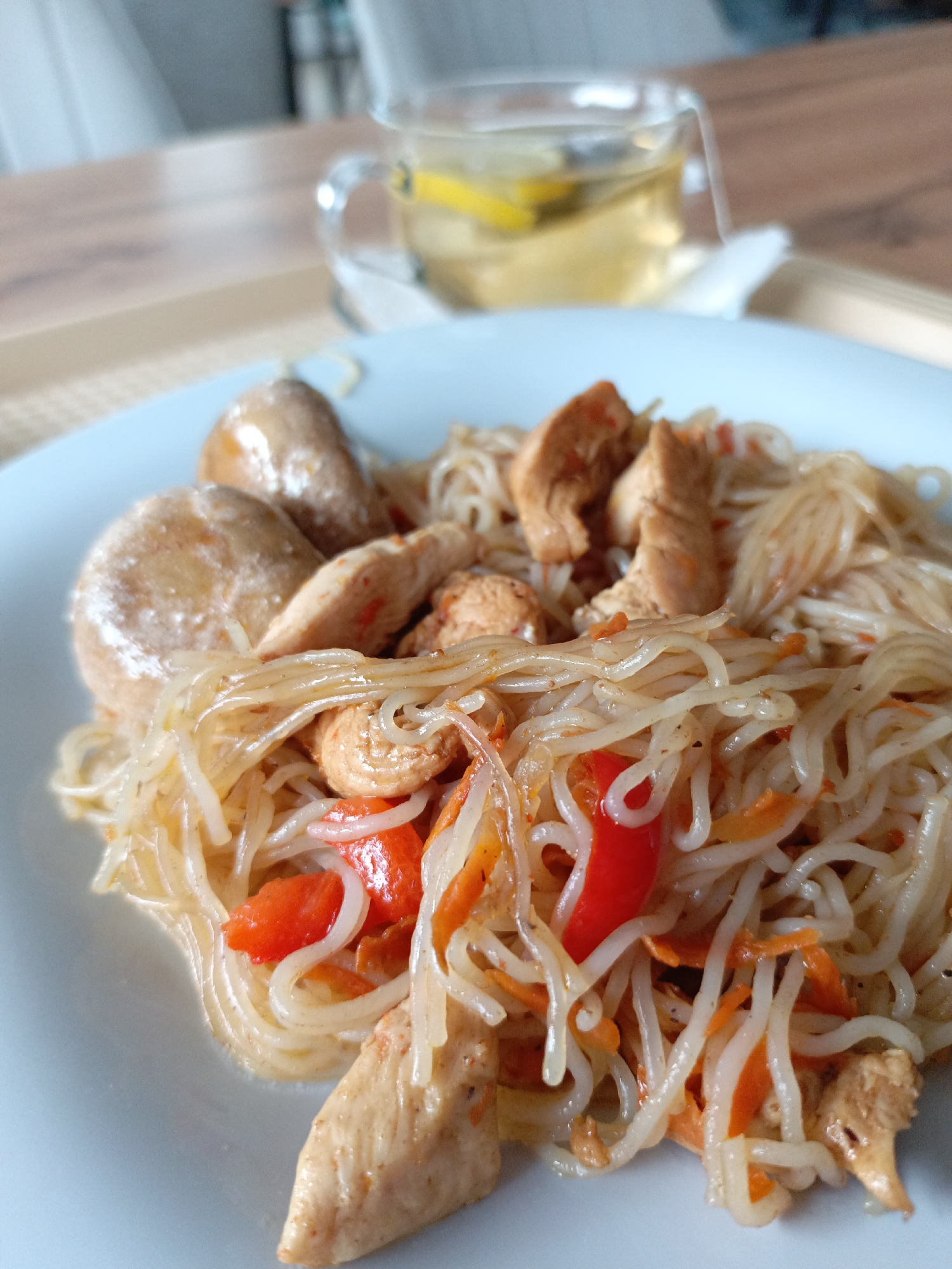 The image displays a meal served on a light-colored placemat over a wood-grained table in an indoor dining setting in the city of Noviy, Russia. In the foreground, a light blue plate holds a dish composed of translucent noodles, irregular chunks of cooked chicken, strips of red bell pepper, and shredded orange carrots. Two prominent, smooth, light-brown, oval-shaped items are positioned on the left side of the plate. In the blurred background, to the right, a clear glass mug with a handle contains a light yellow beverage, identified as tea, with multiple lemon slices and a visible white teabag string. No individuals are present in the frame.