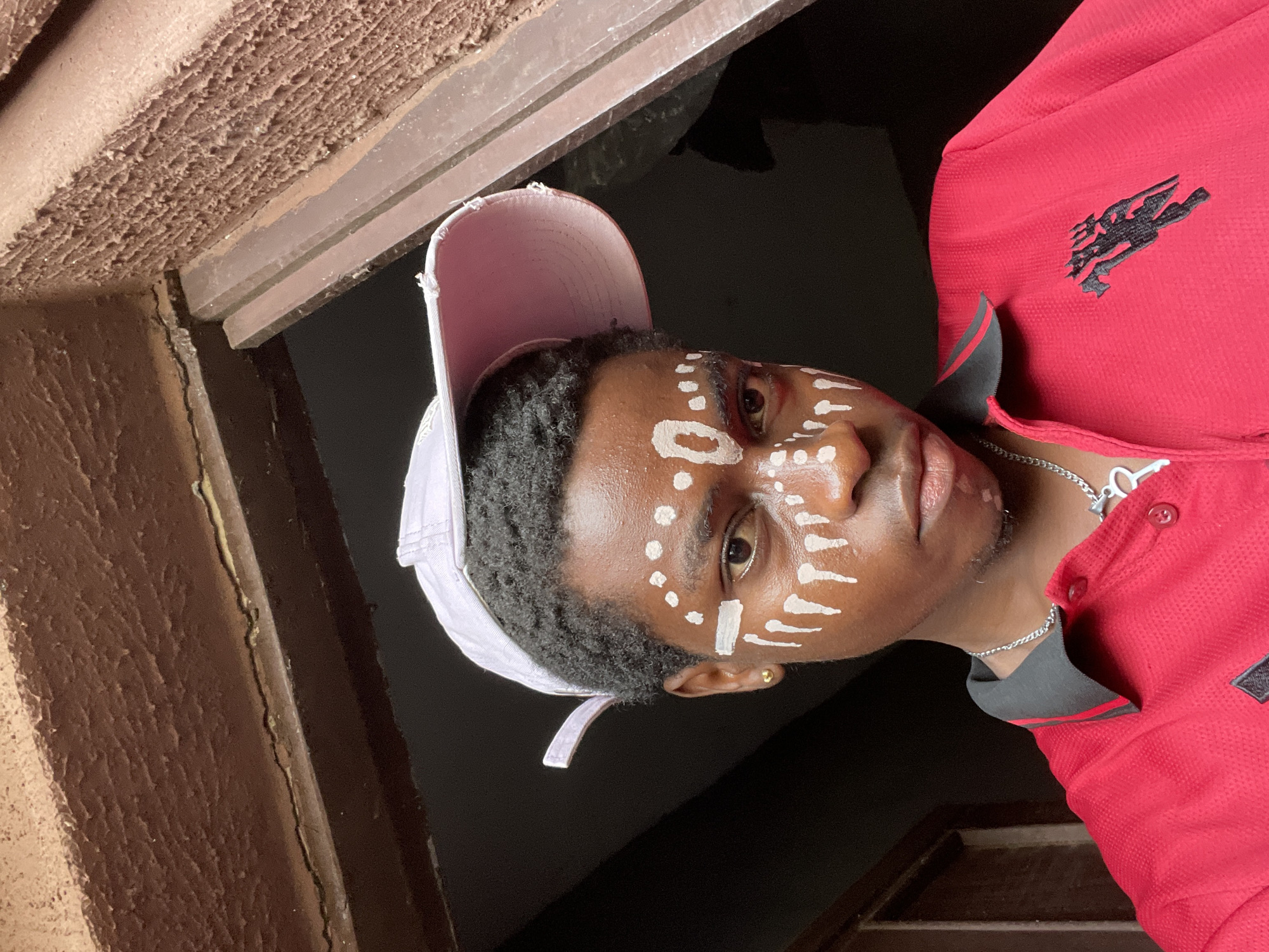A young African man with white paint designs on his face stands in the city of Nkpor, Nigeria. He wears a pink baseball cap and a red polo shirt with a dark grey collar and a black logo on the chest. A silver necklace adorns his neck. The background is dark, suggesting an indoor location, with glimpses of brown wall and trim. The orientation of the photograph is rotated, making the scene appear sideways.