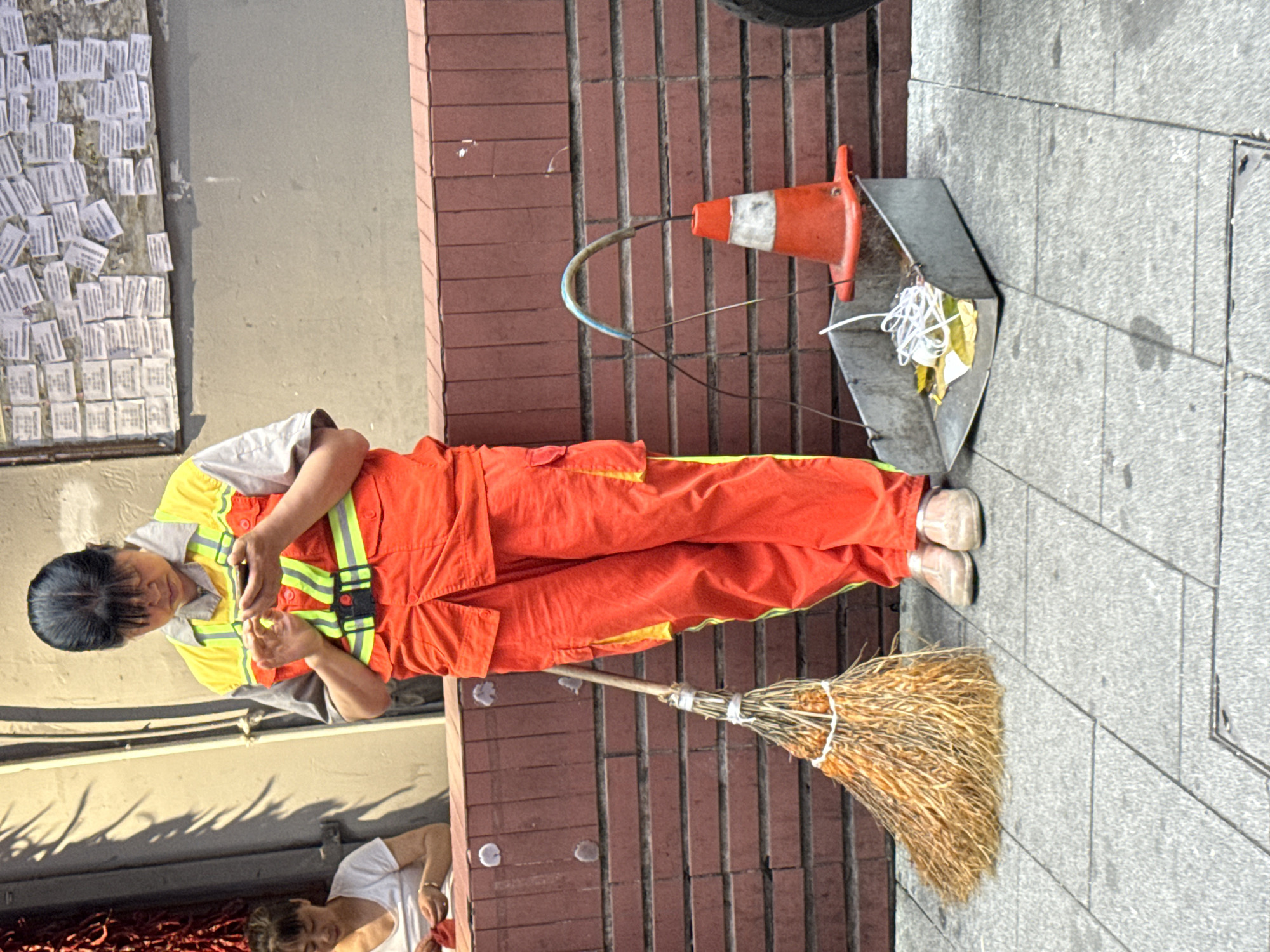 A street cleaner in an orange jumpsuit and yellow reflective vest stands on a gray sidewalk next to a red brick wall in Yuzhong District, China. She is holding a smartphone and looking at it. Beside her is a straw broom, a metal dustpan containing trash, and an orange traffic cone. In the background, there is a wall covered with small papers and another person in a white shirt is partially visible.