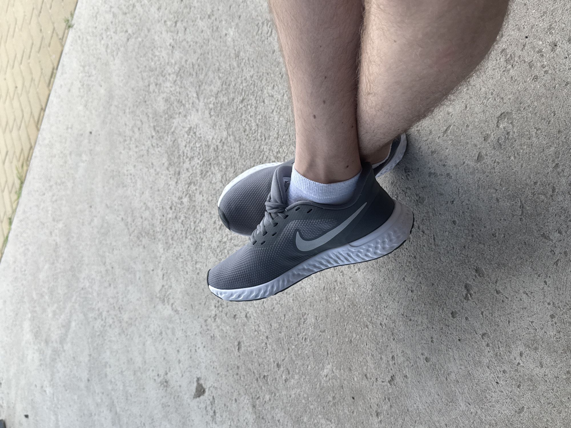 A pair of gray Nike running shoes with white soles and light gray socks are worn by a person whose legs are crossed. The shoes are positioned on a concrete surface that appears to be a sidewalk or pathway. The background consists of more concrete and a section of yellow-toned paving stones. The image provides a close-up view of the lower legs and feet, with the rest of the person's body not visible. The scene is outdoors, suggesting a casual setting.