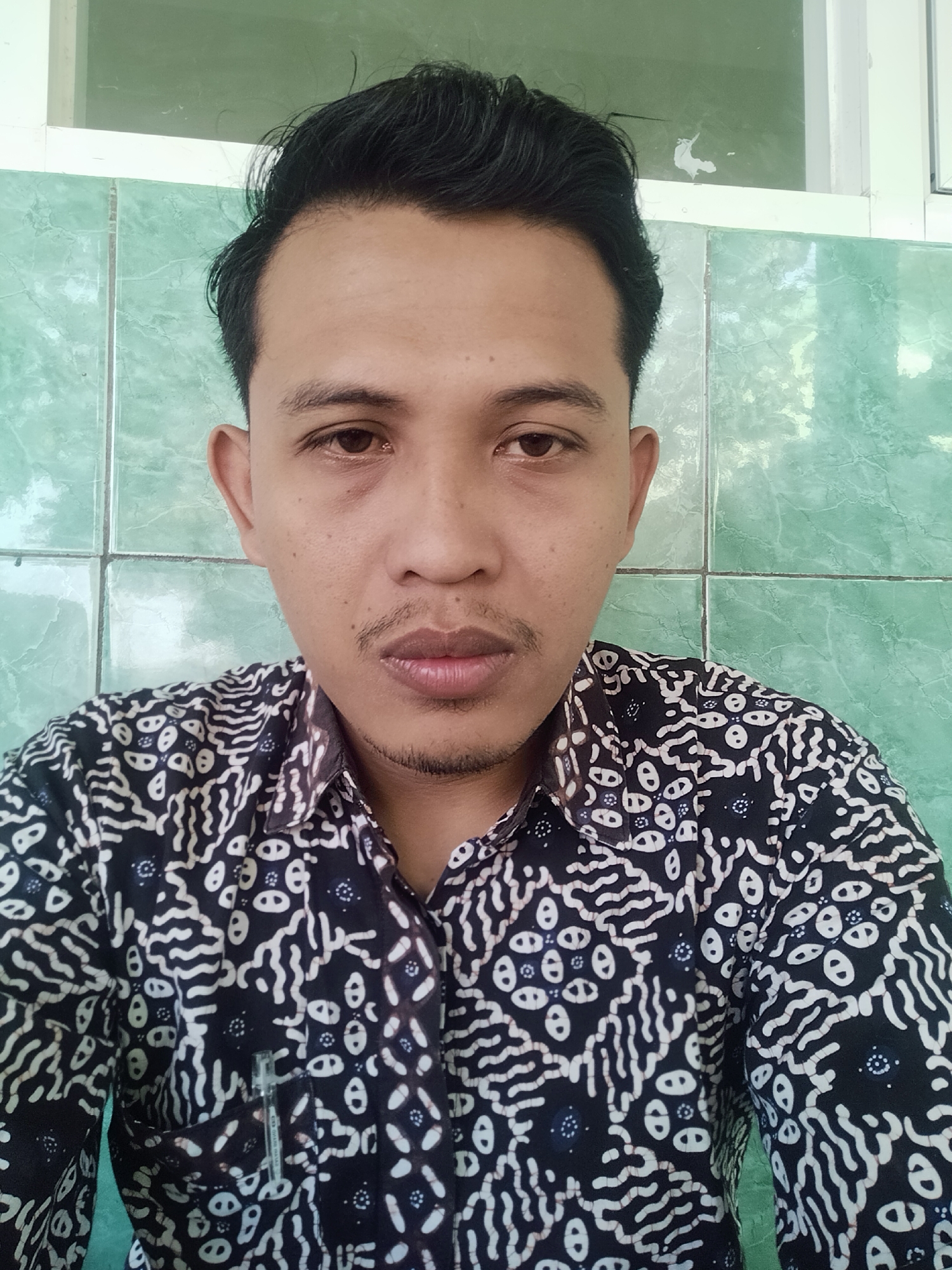 A man is shown from the chest up, facing the camera, in what appears to be an outdoor or semi-outdoor setting. He has dark hair styled to the side, a thin mustache, and a goatee. He is wearing a dark button-up shirt with a white batik pattern. Behind him, the wall is tiled with pale green squares. The setting is likely a residential area or a simple public building in Pucakwangi, Indonesia.
