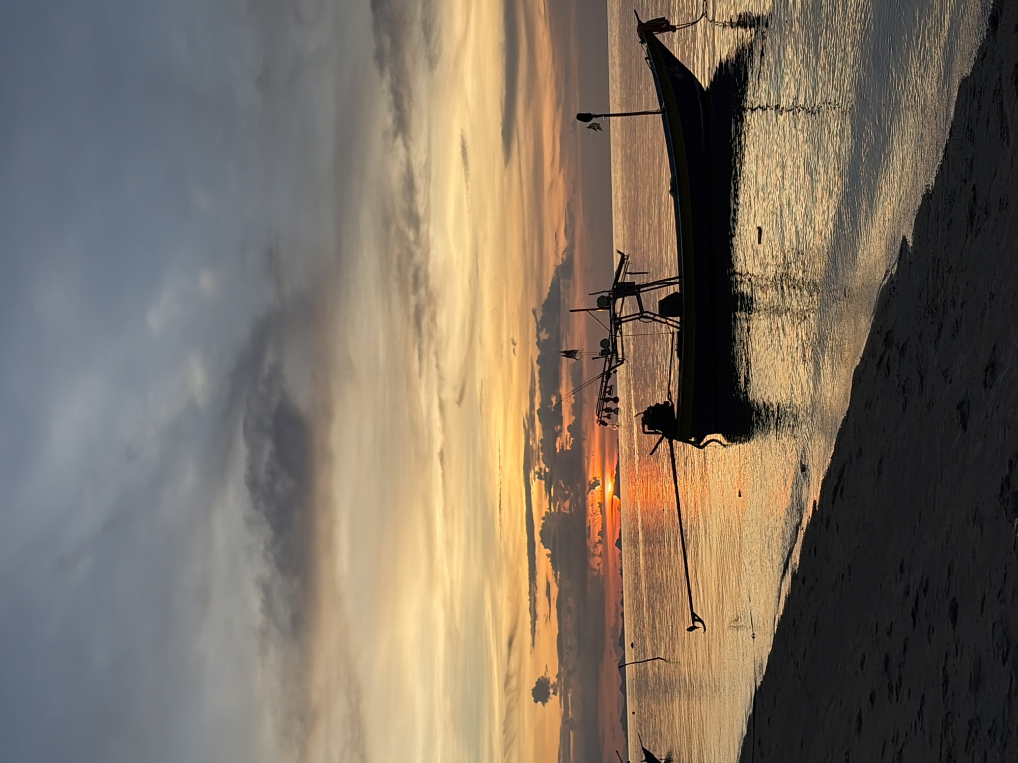 A tranquil scene unfolds at Baan Thong Po, Thailand, as the sun dips below the horizon, casting a warm, golden glow across the sky and water. The sky is a canvas of soft oranges, yellows, and dusky grays, with scattered clouds adding texture to the twilight. In the foreground, a sandy beach stretches along the left side of the frame, its dark silhouette contrasting with the illuminated water. The calm sea reflects the vibrant colors of the sunset, creating a shimmering spectacle.
Dominating the mid-ground is a longtail boat, its dark hull silhouetted against the radiant water. The boat is anchored and at rest, its distinctive long propeller shaft extending into the water. A small cabin structure is visible on the boat, along with a few fishing rods or antennas. Another longtail boat is partially visible on the far left edge of the frame, also silhouetted. In the distance, a faint outline of islands or landmasses can be seen against the horizon. The overall atmosphere is one of peace and natural beauty, characteristic of a sunset by the sea in this Thai locale.