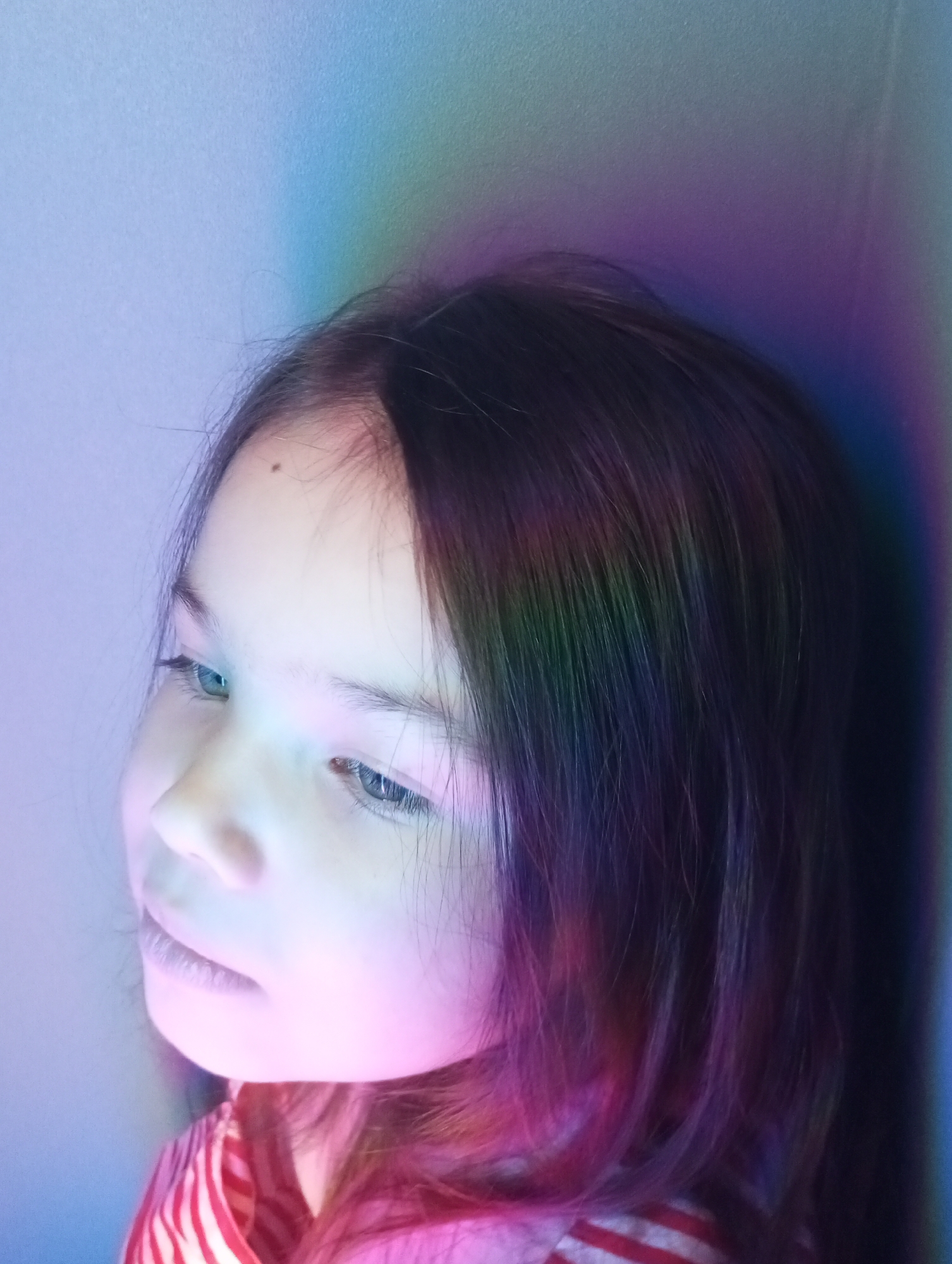 A young girl with dark hair is illuminated by a rainbow of light. Her head is turned slightly to the left. She has a mole on her forehead. She is wearing a striped shirt. The background is a plain wall with the same rainbow light projected onto it. There is no evidence to suggest that the photo was taken in the city of Pervomaysky, Russia.