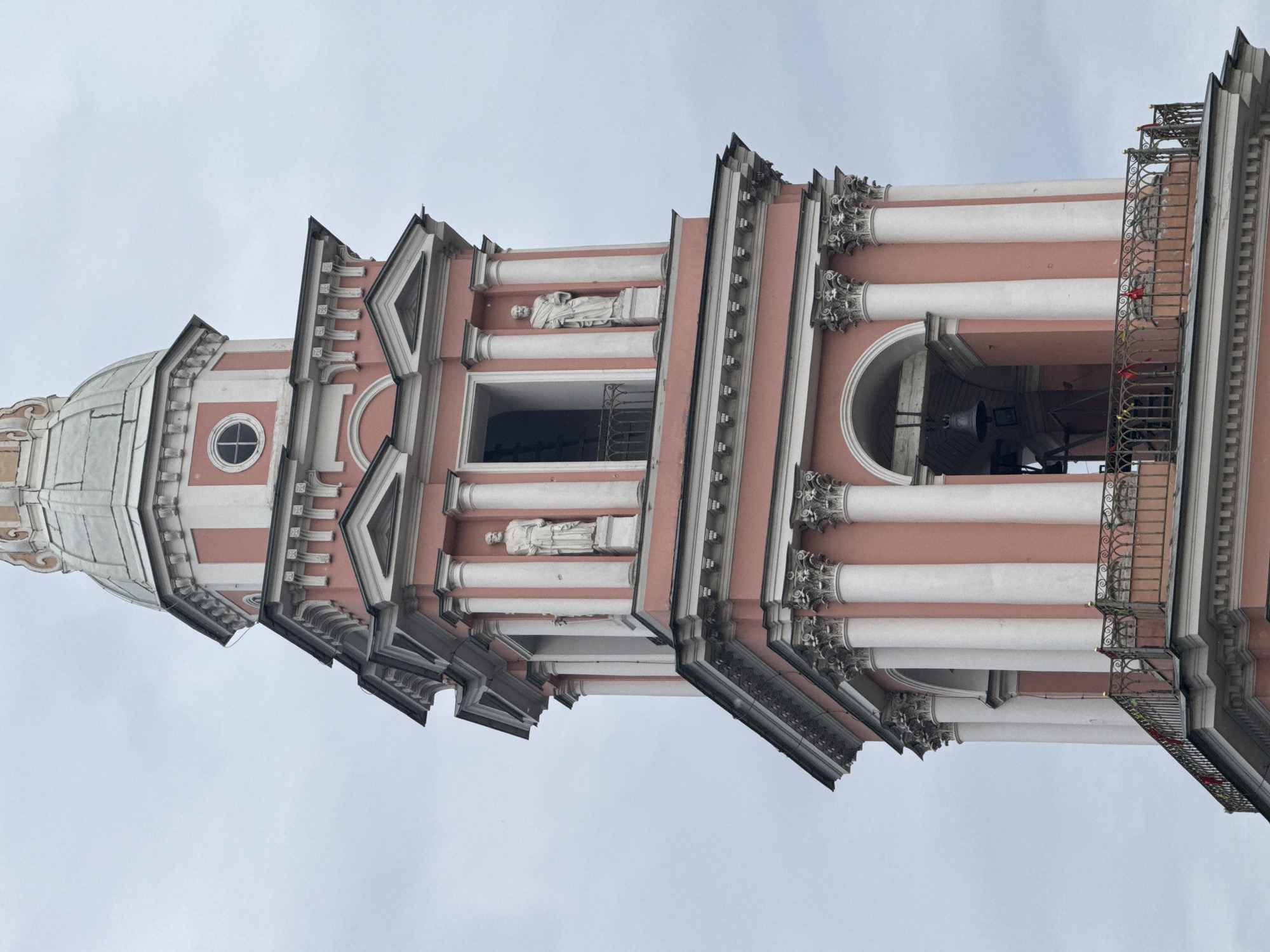 This is a close-up, eye-level shot of the bell tower of the Smolny Cathedral in Saint Petersburg, Russia. The tower is painted a distinctive pink with white architectural details, including columns, cornices, and decorative elements. At the top of the visible portion of the tower, a bell is hanging within an arched opening. Two statues of men in flowing robes stand in niches on either side of an upper window. The tower is topped with a domed roof. A decorative metal railing with red ribbons tied to it runs along a balcony. The sky is overcast and a pale gray.