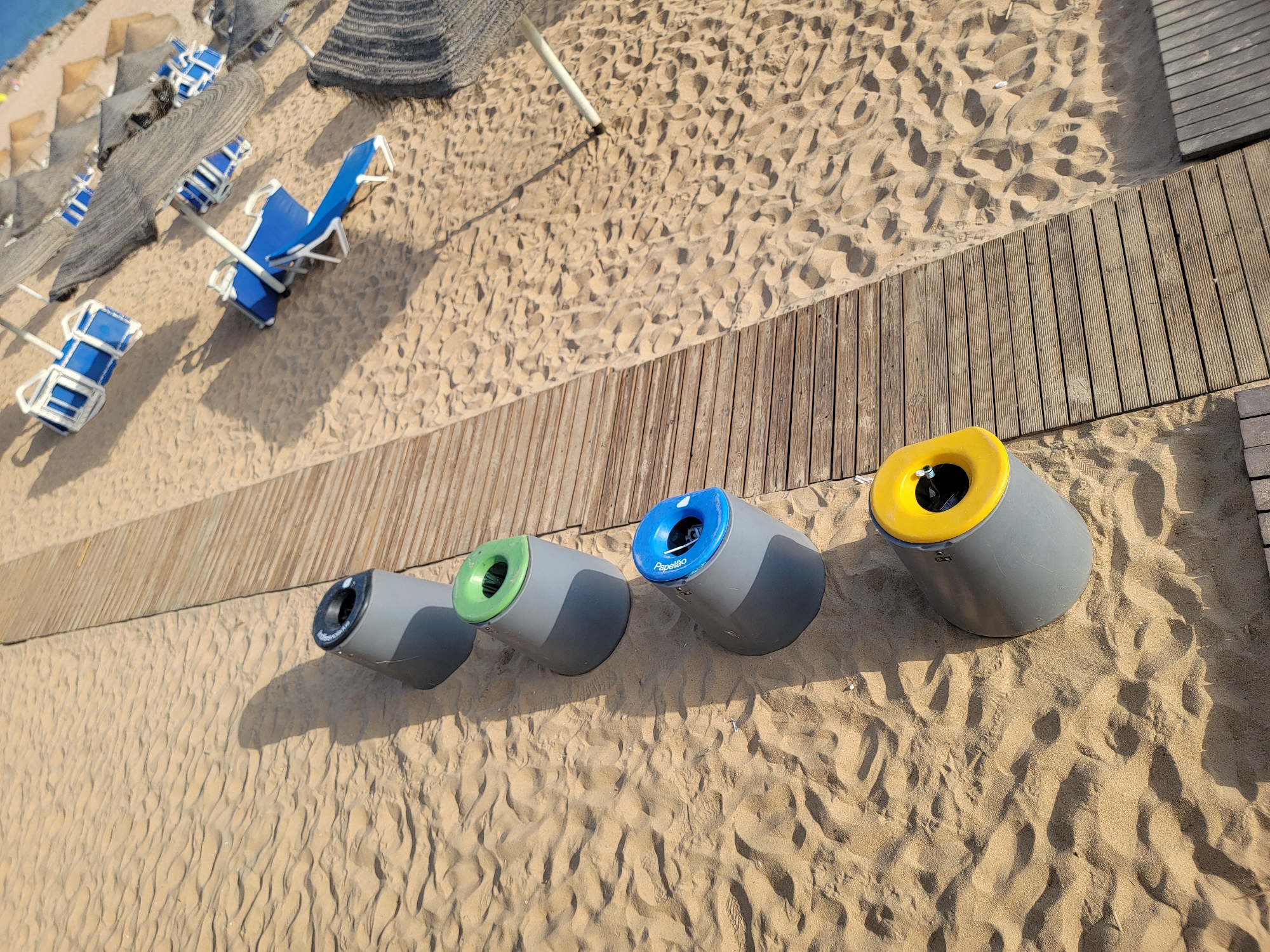 The image shows a beach scene in Quarteira, Portugal. In the foreground, four cylindrical recycling bins are lined up on the sand, each with a colored lid: black, green, blue, and yellow. The blue lid has the word "Papelao" written on it, indicating "cardboard" in Portuguese. The bins are placed next to a wooden boardwalk. In the background, beach umbrellas and lounge chairs are scattered on the sand, suggesting a sunny day at the beach. The sand is imprinted with footprints. The lighting and shadows indicate it is daytime.