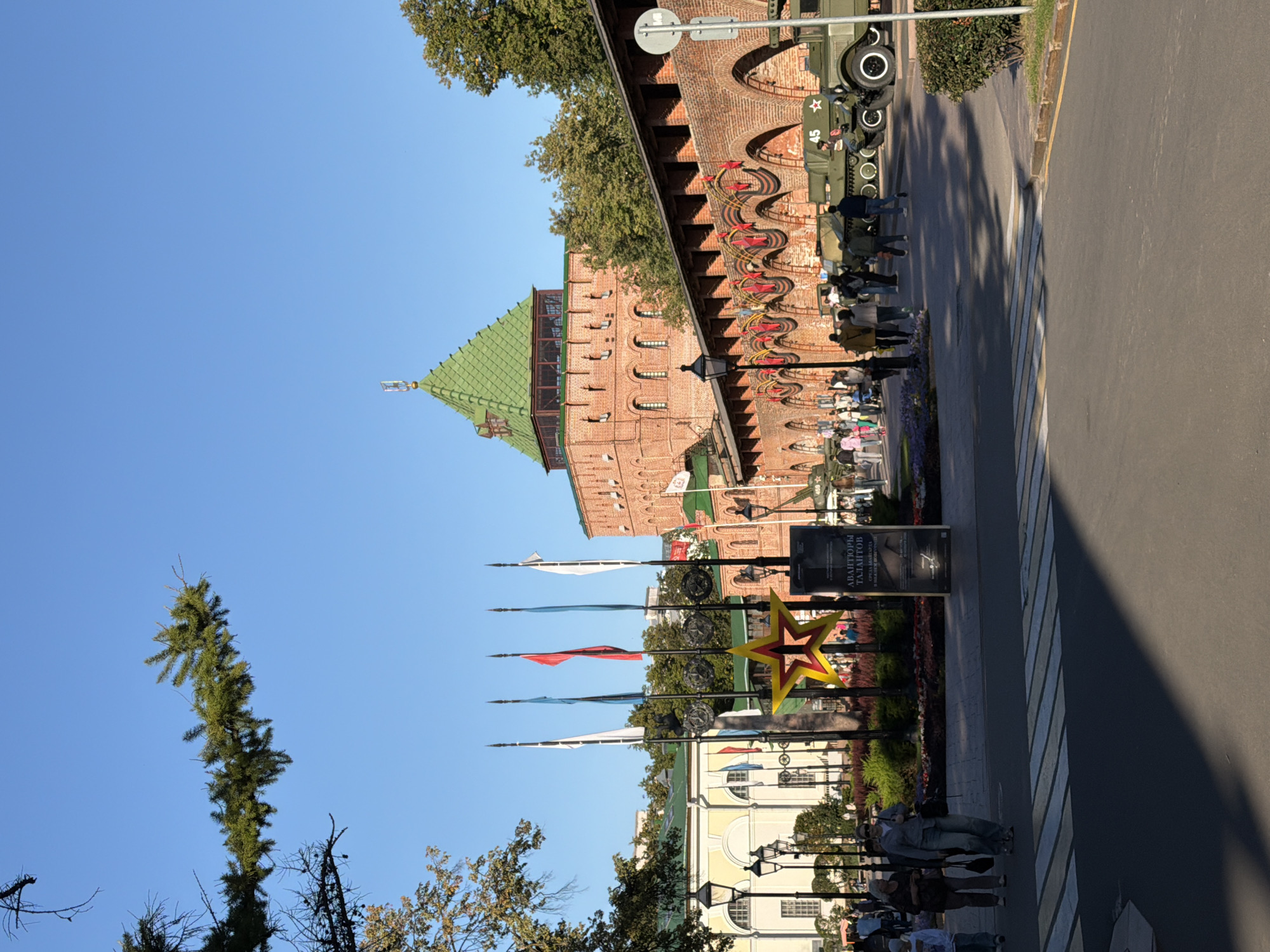 The image depicts a view of the Nizhny Novgorod Kremlin in Nizhny Novgorod, Russia. In the foreground, a paved road with pedestrian crossings is visible. To the left, several people are walking, and to the right, military vehicles are displayed.
The middle ground features a section of the Kremlin wall, constructed of red brick, with arched openings. Red ribbon decorations are hung along the top of the wall, interspersed with red star emblems. A large, green-roofed brick tower, a prominent feature of the Kremlin, stands further back on the left side. In front of the tower and wall, there are flags on poles, and a large yellow star decoration is displayed. A sign with Russian text is also present. Beyond the Kremlin wall and tower, trees and other buildings can be seen.
The background is dominated by a clear, bright blue sky. Sunlight illuminates the scene, casting shadows on the road. The overall impression is a bright, sunny day with a historical and commemorative atmosphere.