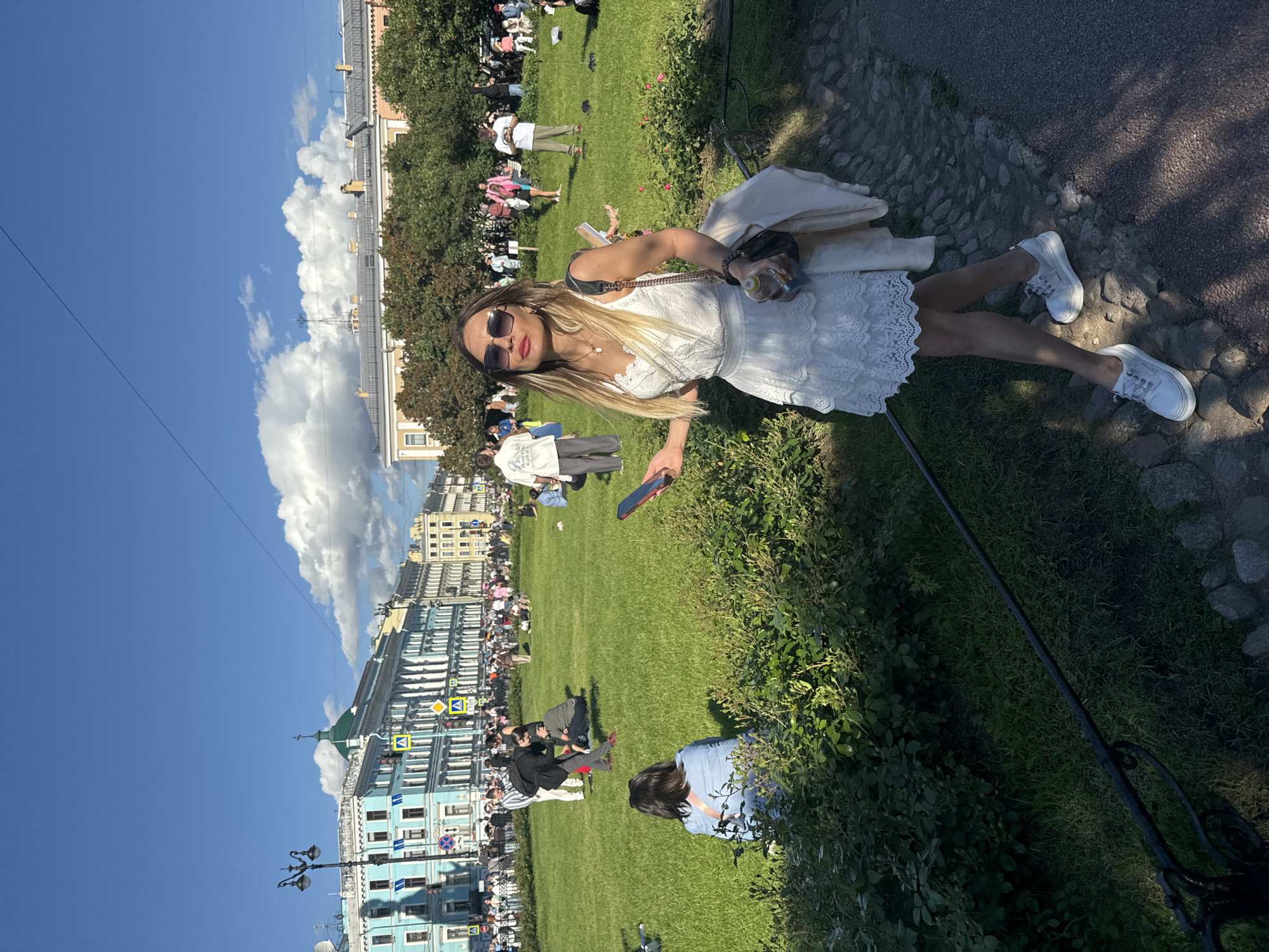 This is a full-body, eye-level shot of a woman in a white lace dress standing on a cobblestone path in front of a large grassy park in Saint Petersburg, Russia. The woman is facing the camera, with her left hand holding a smartphone and her right hand near her hip. She is wearing sunglasses and has blonde hair. A light-colored jacket is draped over her right shoulder, and she carries a black bag. Behind her, the park is populated with many people enjoying the sunny day. Some are sitting on the grass, while others are walking or standing. In the background, there are several historical buildings with distinct architectural features, characteristic of Saint Petersburg. The sky is blue with fluffy white clouds, indicating good weather. The image appears to have been taken in the afternoon. The overall scene depicts a leisurely day in a public urban space in Saint Petersburg.