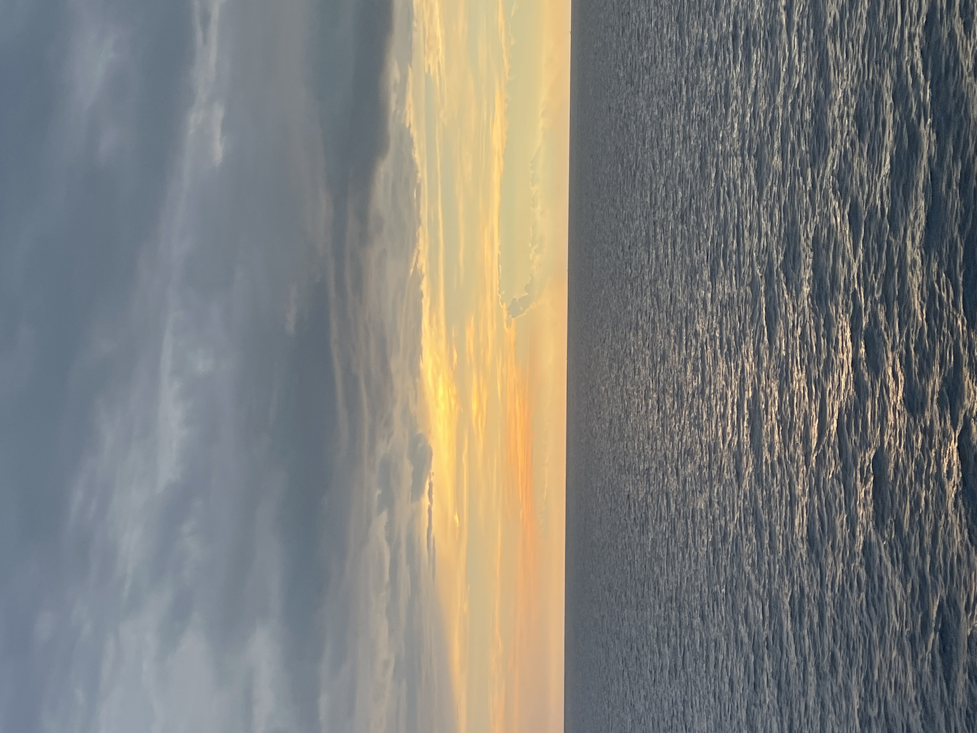 This is a vertical, eye-level, medium shot of a calm sea and sky during sunset. The image is split vertically, with the left half depicting the sky and the right half showing the sea. The sky is filled with clouds, some dark gray and others tinged with warm orange and yellow hues from the setting sun. The clouds are streaky and wispy, with a few patches of bright sunlight breaking through. The sea is a deep blue, with gentle ripples creating a textured surface. The sunlight reflects off the water, creating a shimmering effect. The horizon line is sharp and clear, dividing the sky and sea. The overall mood of the image is peaceful and serene, evoking a sense of tranquility. The time of day is clearly sunset, as indicated by the warm colors in the sky and the low angle of the sun. The weather appears to be partly cloudy. The location is indeterminate, but the presence of a vast expanse of water suggests a coastal or oceanic setting. There is no visible text in the image.