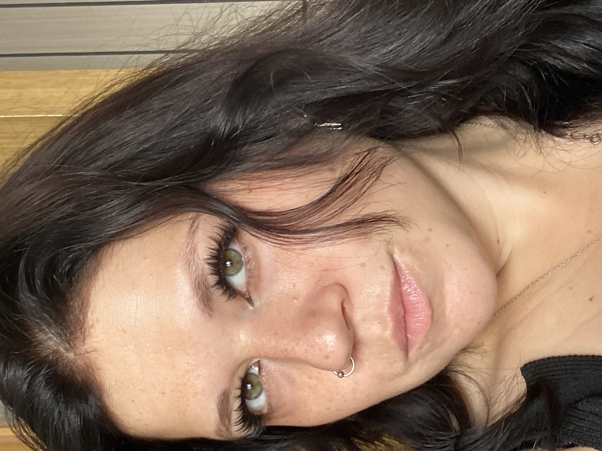 A close-up, eye-level, medium shot captures the right side of a woman's face and upper chest. She has dark, wavy hair, light green eyes with long, dark eyelashes, and fair skin with freckles. A silver hoop earring is visible in her right nostril. She wears a black top and a silver necklace with a pendant. The background consists of vertical wooden blinds. The image does not provide any information about its location in the city of Unknown, Russia.