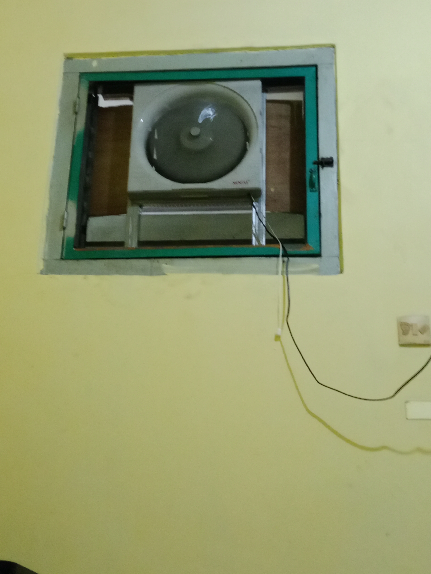 The image shows a window with a fan installed. The window frame is painted green and gray. The fan is a box fan with a gray casing. There are wires coming from the fan and extending down the wall. A light switch is visible on the yellow wall to the right of the window. The environment appears to be indoors, likely a residential or commercial space. The specific location in West Jakarta, Indonesia is not discernible from the image.