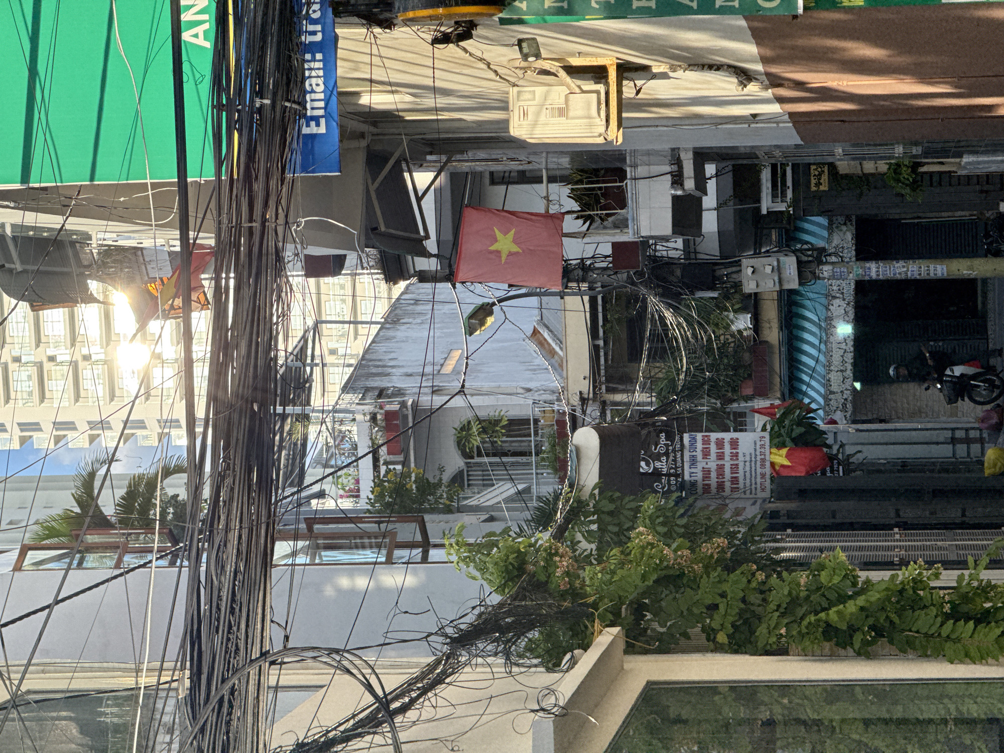 This is a street-level photograph taken in Nha Trang, Vietnam, looking down a narrow alleyway. The scene is characterized by a dense network of overhead electrical wires, a common sight in Vietnamese cities. In the foreground, to the left, is the side of a building with a large window. The alleyway opens up to reveal several stories of buildings on either side, showcasing balconies with potted plants and open windows. A red Vietnamese flag with a yellow star hangs prominently on the right side of the alley. Sunlight streams down from the upper right, creating bright highlights and casting shadows. On the right side of the alley, there is an air conditioning unit mounted on the wall. Further down the alley, a person on a motorcycle is visible, partially obscured by shadows. Signs with Vietnamese text are present on the buildings, indicating businesses. The overall impression is a vibrant, urban environment with a sense of verticality created by the multi-story buildings and the tangled wires overhead.