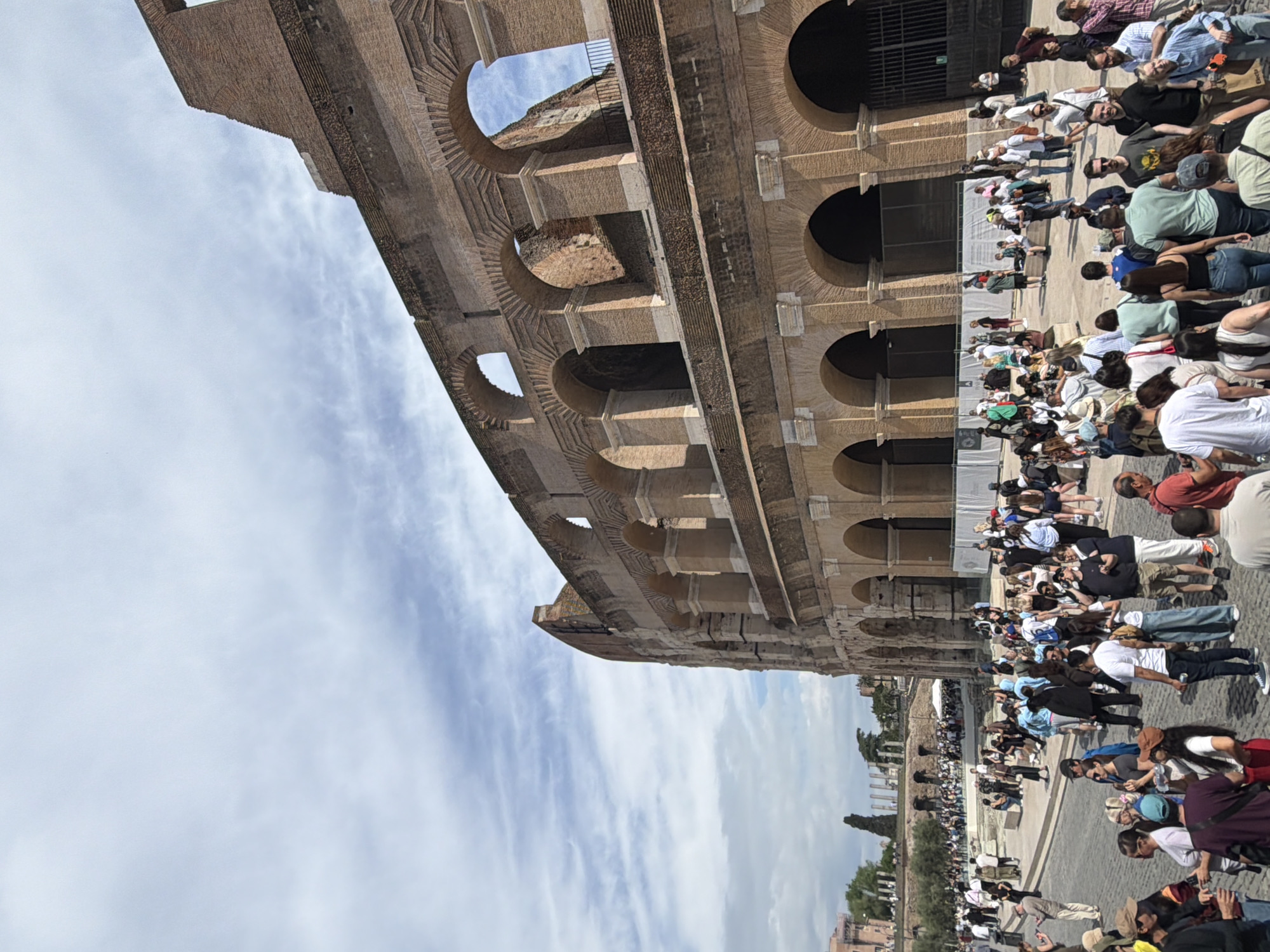 The image displays a significant portion of the Colosseum in Rome, Italy, under a bright, partly cloudy sky. The monumental ancient amphitheater, built of brick and stone, occupies the left and central areas of the frame, showcasing its iconic tiered arcades and numerous arched openings. Portions of the inner structure and weathered stone are visible through the exterior arches.
In the right foreground, a large and diverse crowd of people is gathered, appearing to be tourists or visitors. Individuals are seen standing, walking, and forming lines, suggesting they are either waiting to enter the Colosseum or observing it from the exterior. They are dressed in various casual attires, indicative of a warm, sunny day. Some are interacting in groups, while others are looking towards the historical structure.
The sky above is a light blue with scattered white, wispy clouds, indicating clear daytime weather, likely late morning or early afternoon. The scene conveys a busy atmosphere typical of a popular tourist attraction. In the distant background, to the far right, faint outlines of trees and other ancient Roman structures can be discerned. No discernible text is visible on the Colosseum itself or among the crowd.