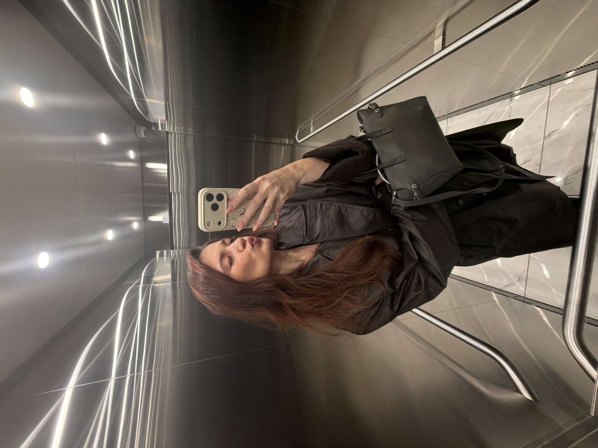 A person with long, auburn hair is taking a selfie in an elevator with stainless steel walls and linear lights. The person is wearing a black coat and a black handbag is visible. The elevator floor appears to be tiled with a marble-like pattern. The location is likely within a modern building, possibly in Belgrade, Serbia, but there is no way to verify it from the visual information in the image.