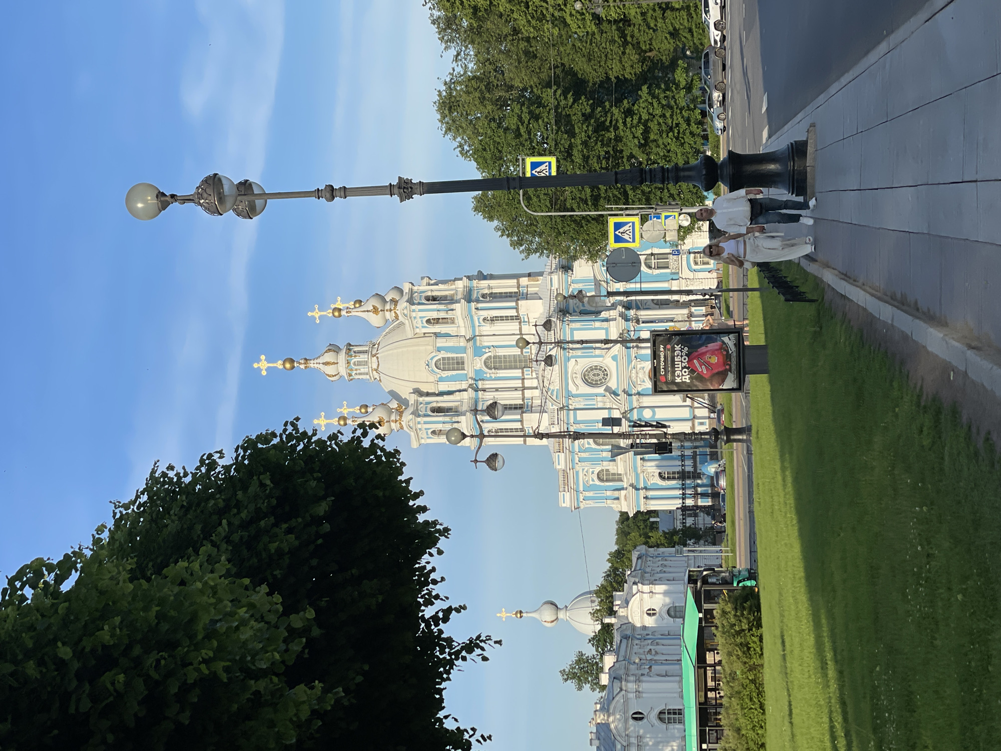 This image captures a street scene in Saint Petersburg, Russia, featuring the Smolny Cathedral and Convent complex under a clear blue sky.
In the foreground, a paved sidewalk runs from the bottom edge towards the mid-ground, separated from a grassy area by a black metal fence. On the sidewalk, right of center, a man and a woman are walking away from the viewer. The woman is wearing a white sleeveless top, white wide-leg pants, and white shoes, with sunglasses perched on her head. The man is wearing a white long-sleeved button-up shirt, dark jeans, white sneakers, and a black fanny pack or belt bag across his chest. A tall, ornate black streetlamp with three globe-shaped light fixtures stands on the sidewalk directly in front of them, extending into the upper right quadrant of the image. Another similar streetlamp is visible further down the sidewalk. To the left of the sidewalk, a lush green grassy area occupies the lower left quadrant, with a green bus partially visible behind a hedge, driving on an unseen road.
In the mid-ground, the prominent Smolny Cathedral dominates the central and right portions of the image. The cathedral is a Baroque structure, distinguished by its white walls with light blue decorative elements and multiple golden onion domes, some topped with crosses. To the left of the cathedral, several white, classical-style buildings with arched windows and pilasters, part of the Smolny Convent, are visible. Further to the left, a smaller white dome with a golden cross atop it rises above the other buildings. A large green tree with dense foliage occupies the upper left corner of the image. A road runs horizontally across the mid-ground, with several cars parked or driving on its right side. Two yellow square signs depicting a pedestrian crossing are visible on poles near the road.
The background consists of a clear, light blue sky with faint wisps of white clouds. Sunlight illuminates the scene from the right, casting distinct shadows on the grassy area and the street.