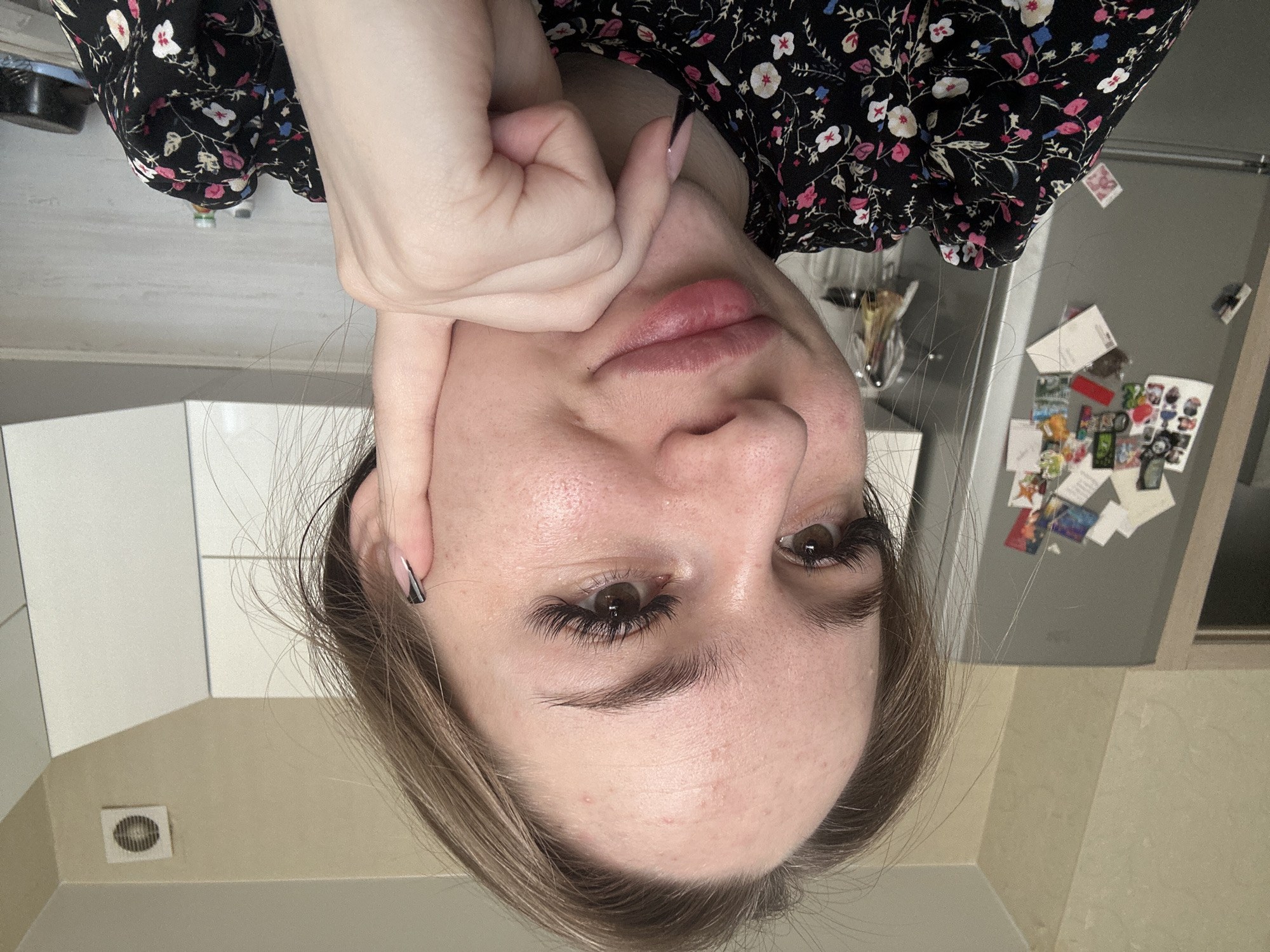 An individual identified as a young adult female is centered in a close-up image, positioned with her head slightly tilted and her left hand resting on her chin. She possesses light brown hair, dark brown eyes, pronounced eyelashes, and full, light-colored lips. Her fingernails are manicured with a two-toned design, primarily black. Her attire consists of a dark-colored garment patterned with white, pink, and blue floral motifs. The immediate background indicates an indoor setting, specifically a kitchen, located in Unknown, Unknown. A metallic-gray refrigerator, covered with numerous decorative magnets and papers, is visible to the left. The walls are painted a neutral light beige. To the right, white overhead kitchen cabinets are present, and a white square ventilation unit with a circular grill is mounted on the wall above them. A light-colored countertop and backsplash are partially visible below the cabinets, displaying various objects.